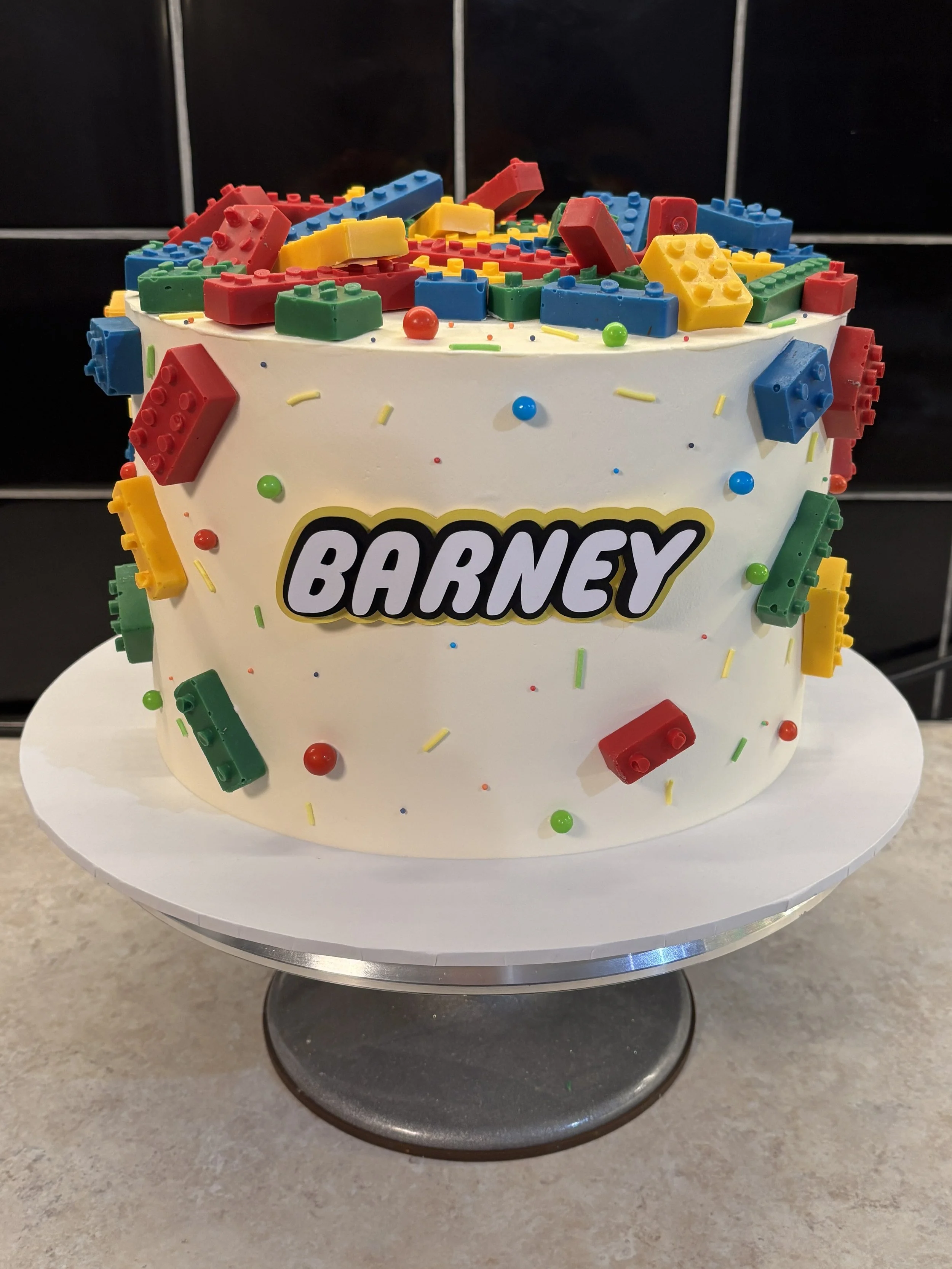 A white birthday cake decorated with colorful Lego bricks and sprinkles, with the name 'Barney' on the front.
