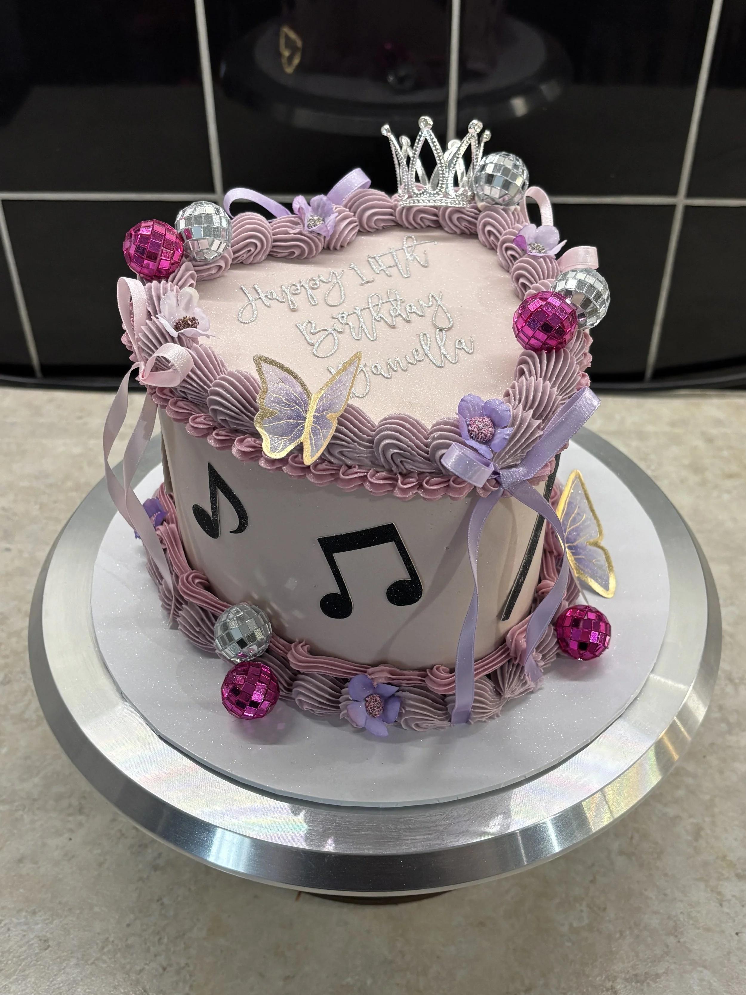 A birthday cake decorated with purple and silver accents, a small crown on top, musical notes, purple flowers, and pink and silver spherical decorations, with the message 'Happy 14th Birthday Ganelia' written on the top.