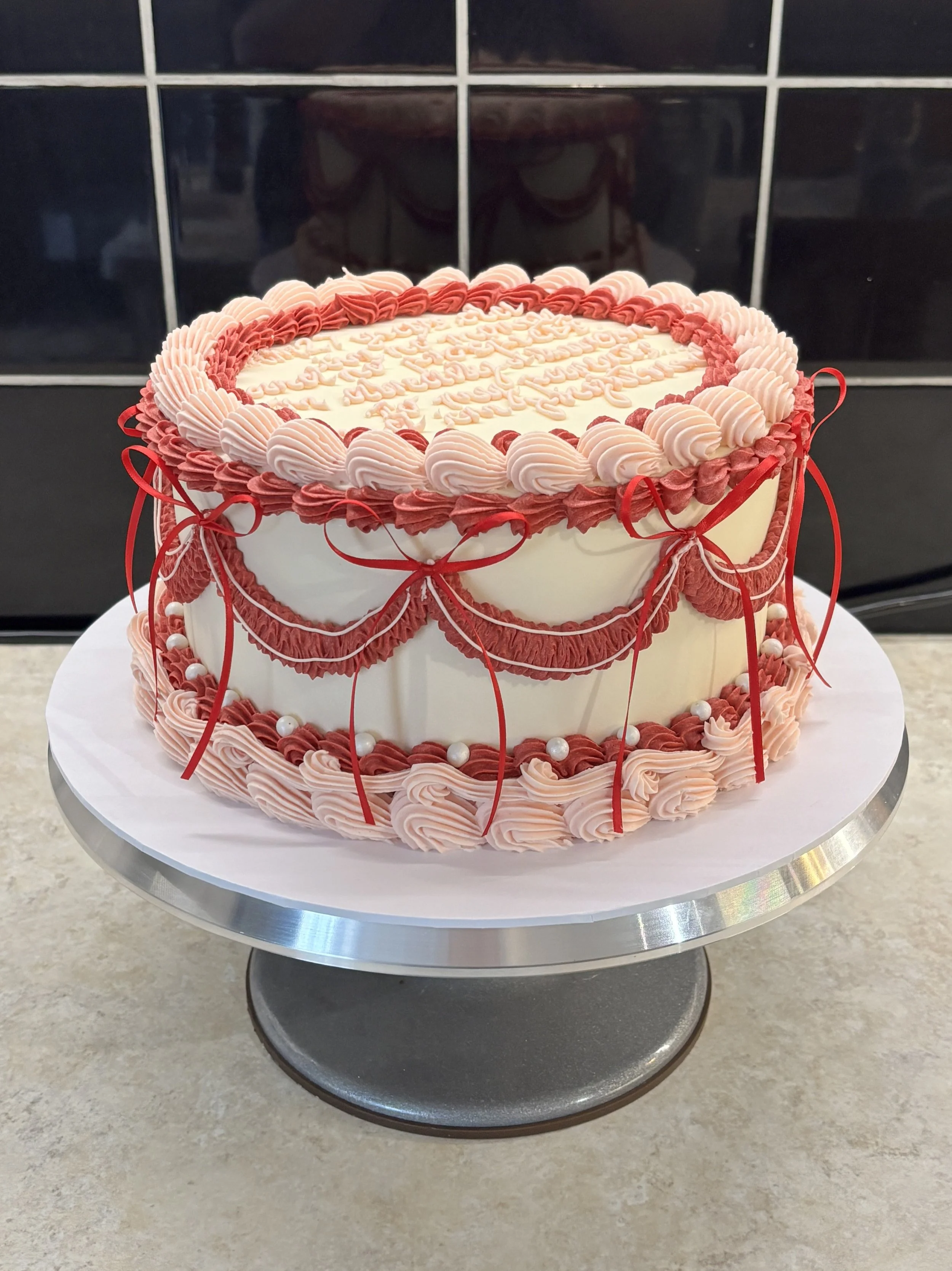 Decorated cake with pink and red icing, bows, and pearl accents on a silver cake stand.