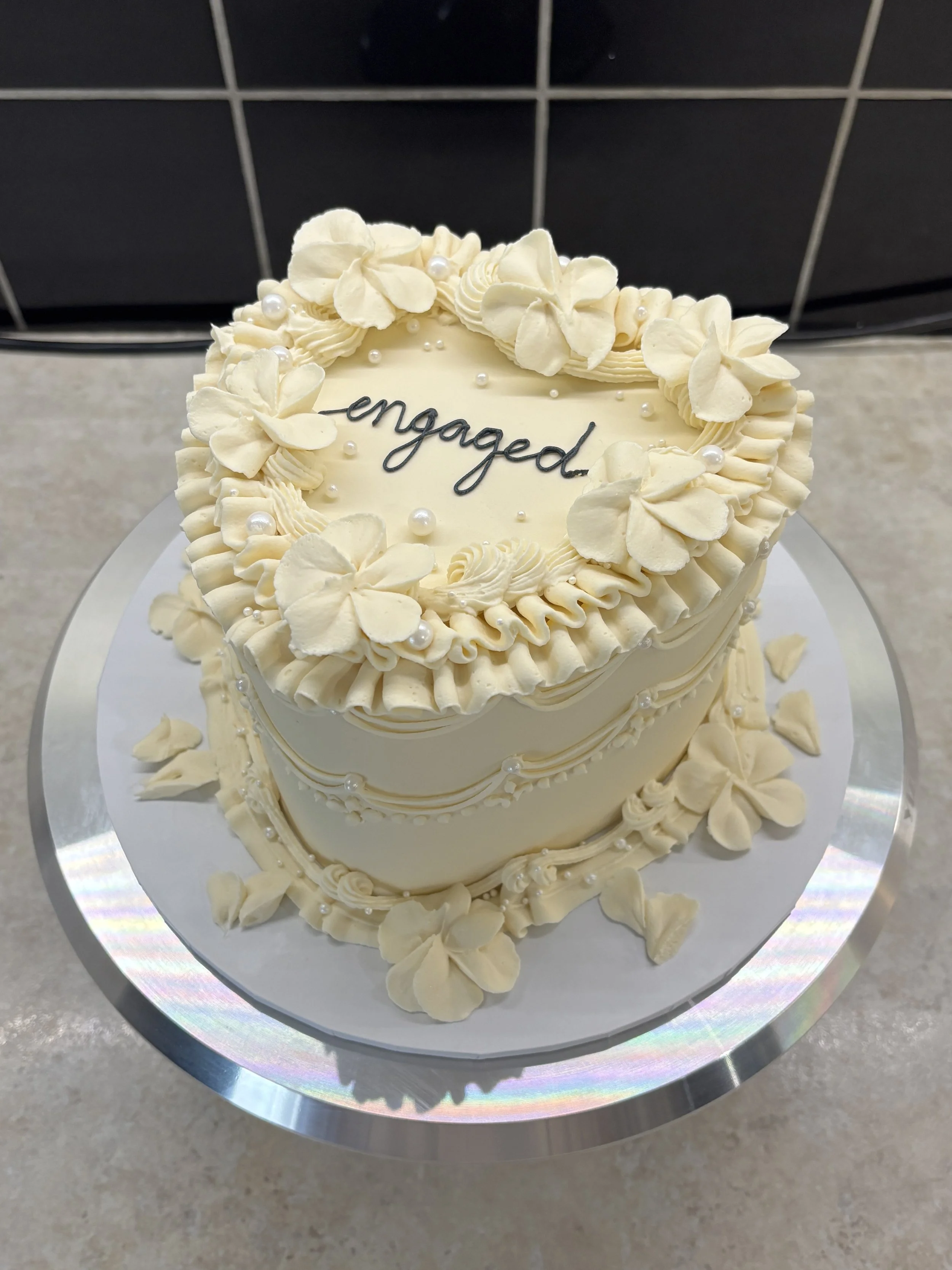 A heart-shaped white cake decorated with white flowers, pearls, and piped frosting, with the word "engaged" written on top.