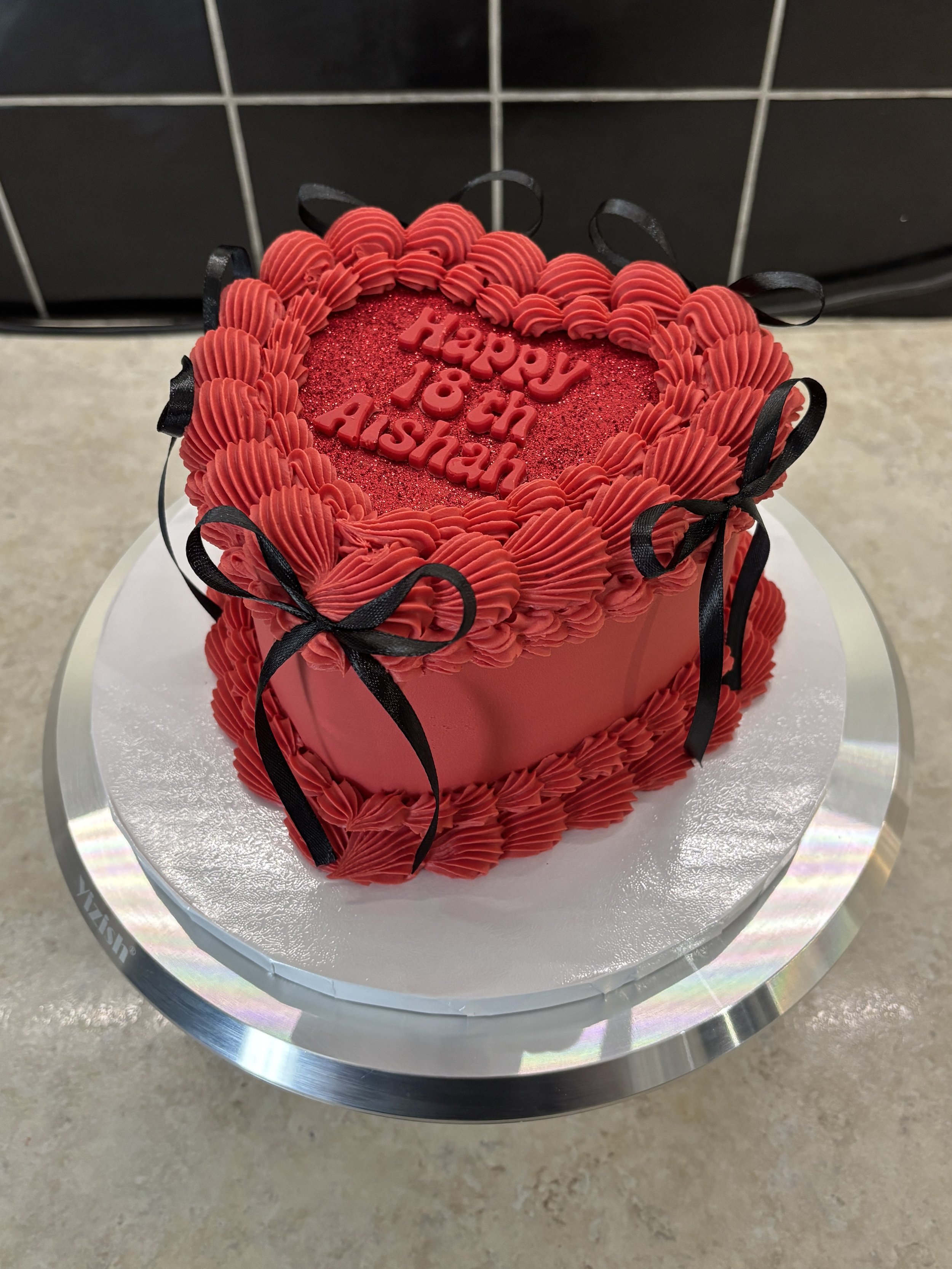 A heart-shaped birthday cake with red frosting and black ribbon decorations, with the message 'Happy 18th Ashan' on top in red lettering.