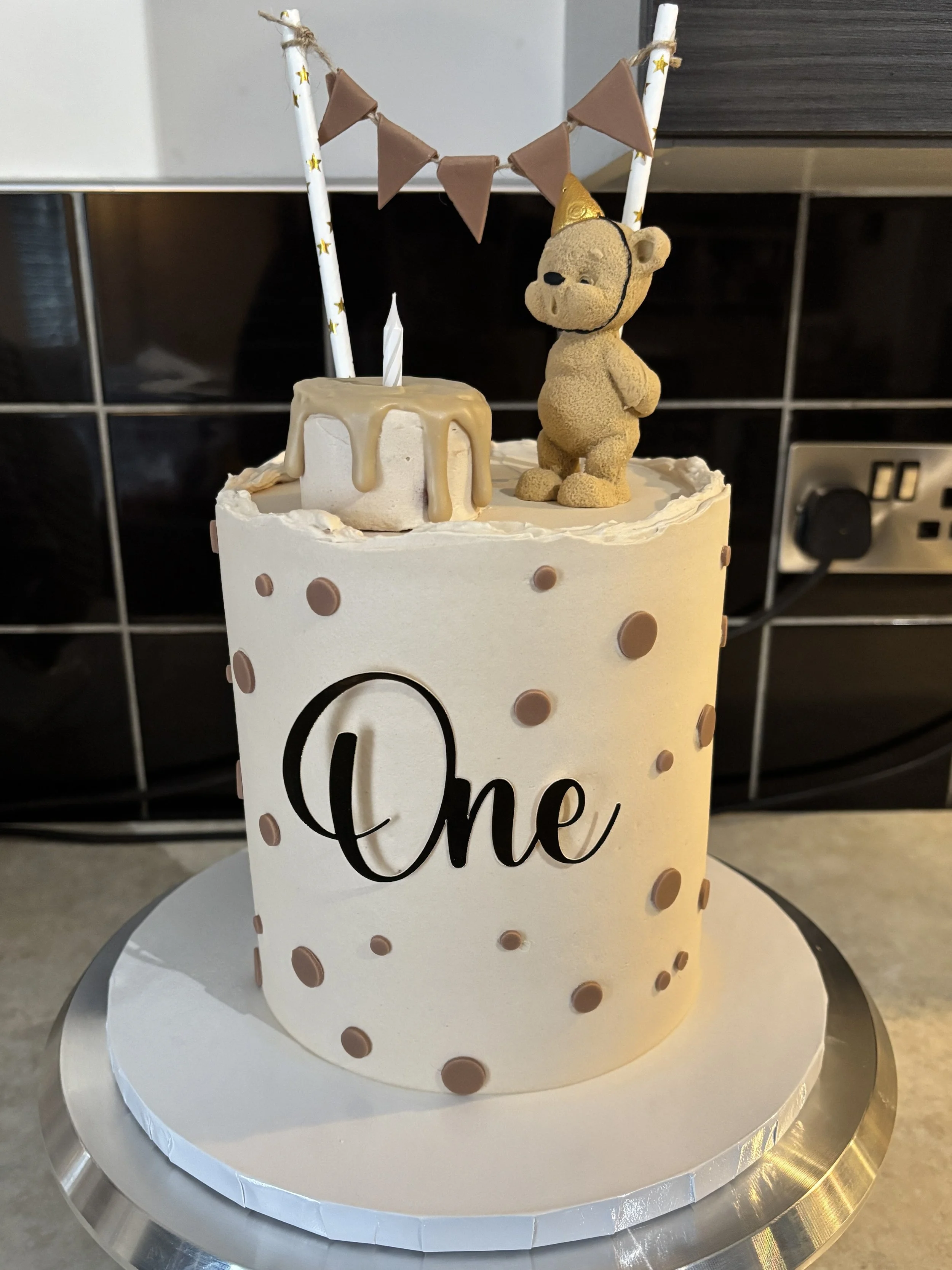 A white birthday cake with chocolate polka dots and the word 'One' in black letters, topped with a small bear figurine wearing a party hat, a candle, and decorative sticks with a bunting banner, on a white cake board.