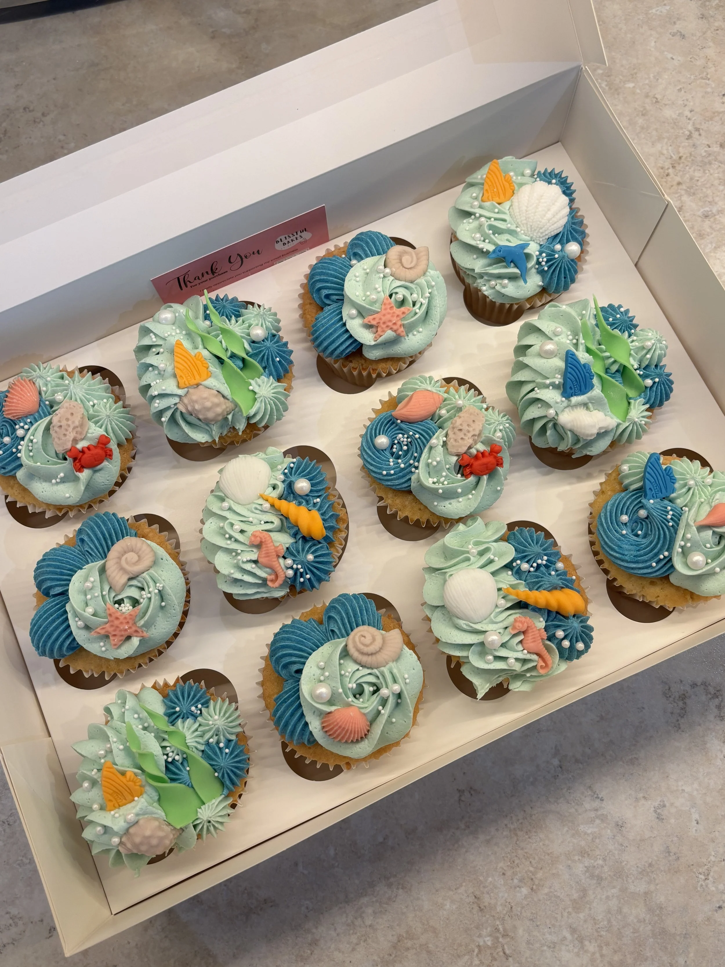 A box of decorated cupcakes with ocean-themed designs, including seashells, starfish, and seaweed, featuring blue, green, and coral-colored frosting.