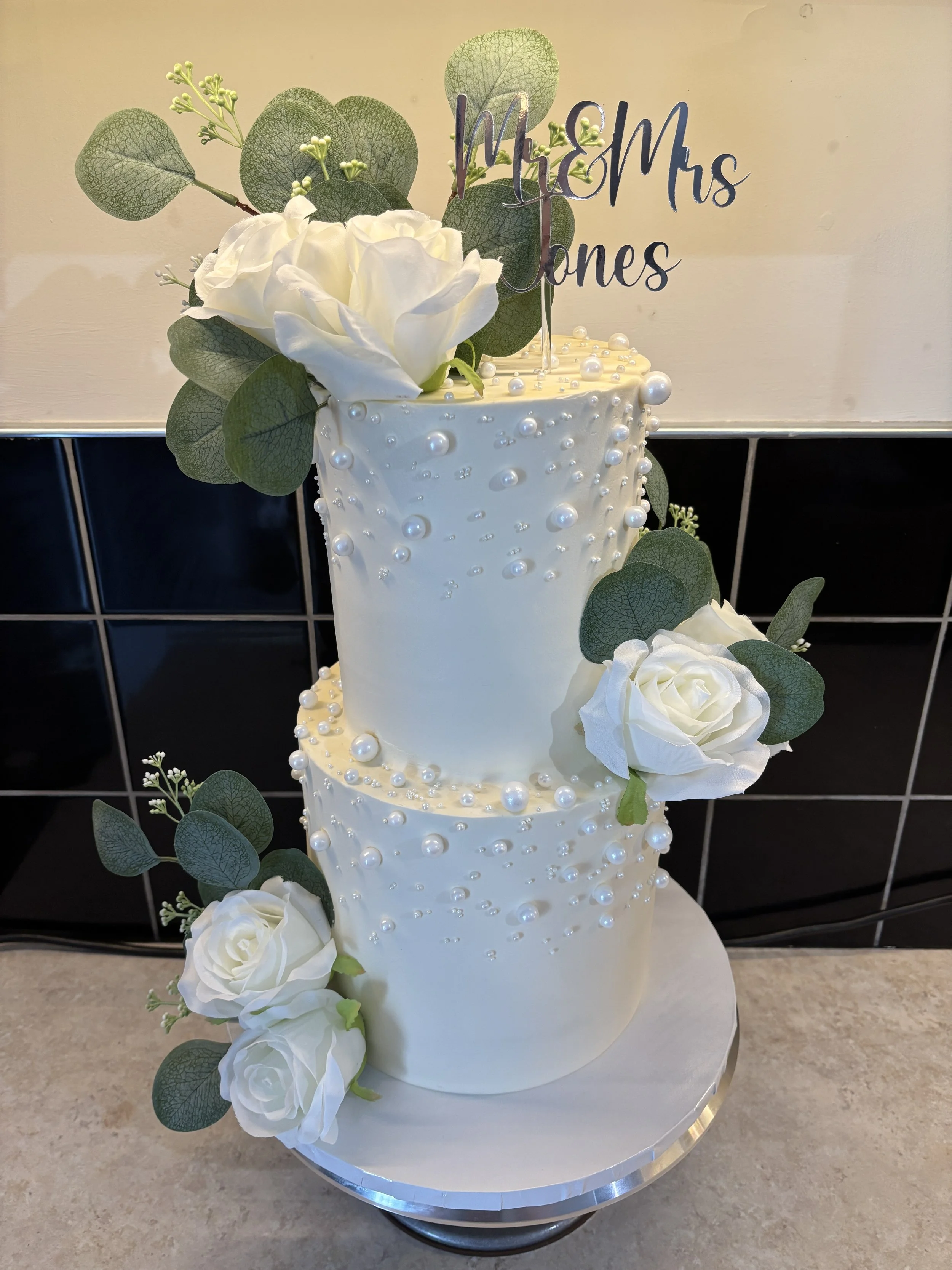 Two-tiered white wedding cake decorated with white roses, green leaves, and pearl-like edible decorations, with a 'Mr & Mrs Jones' cake topper.