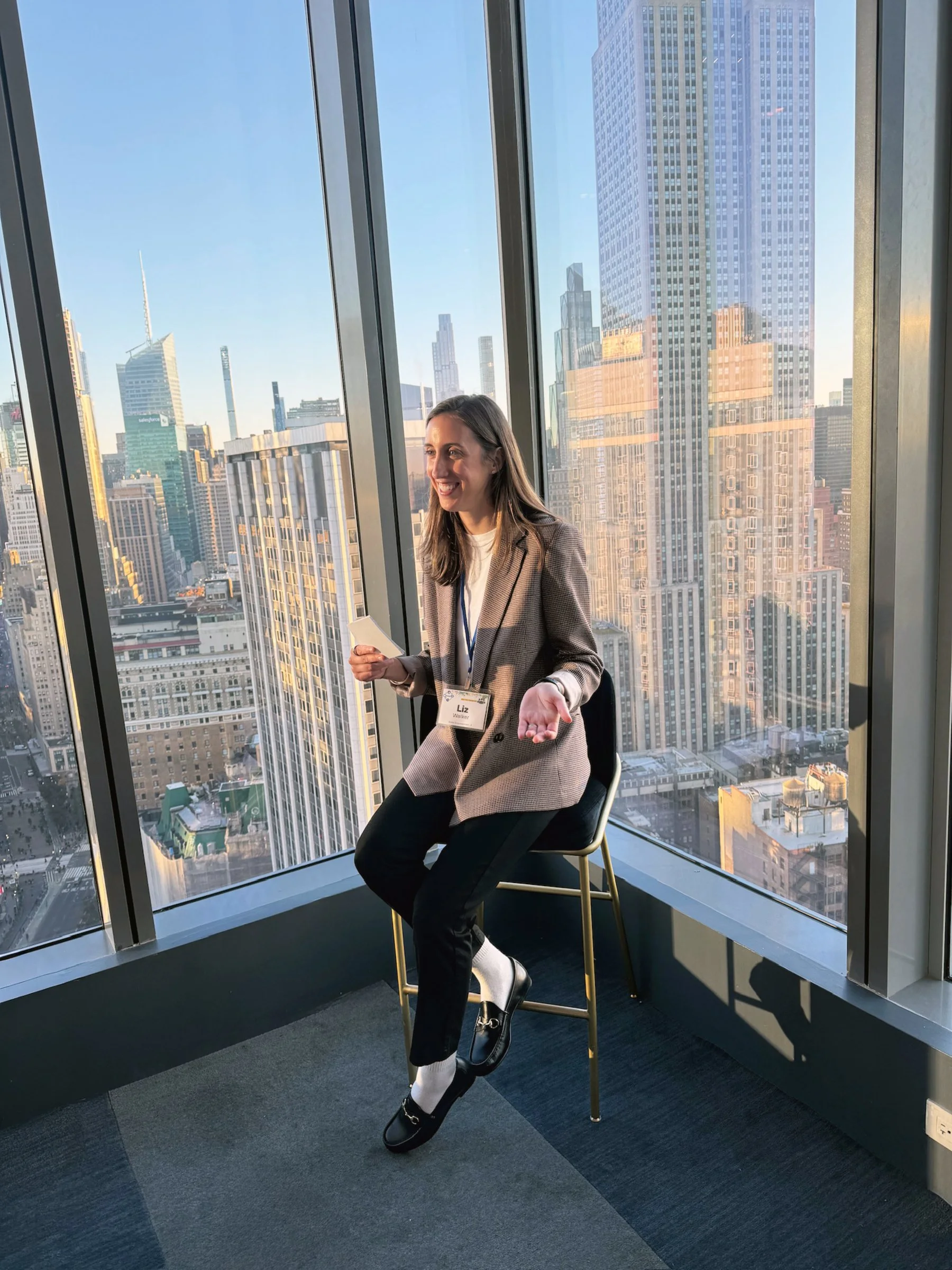 A woman with long brown hair, wearing a checkered blazer, black pants, and black shoes, sitting on a high stool in an office with large windows overlooking a city skyline with tall buildings.