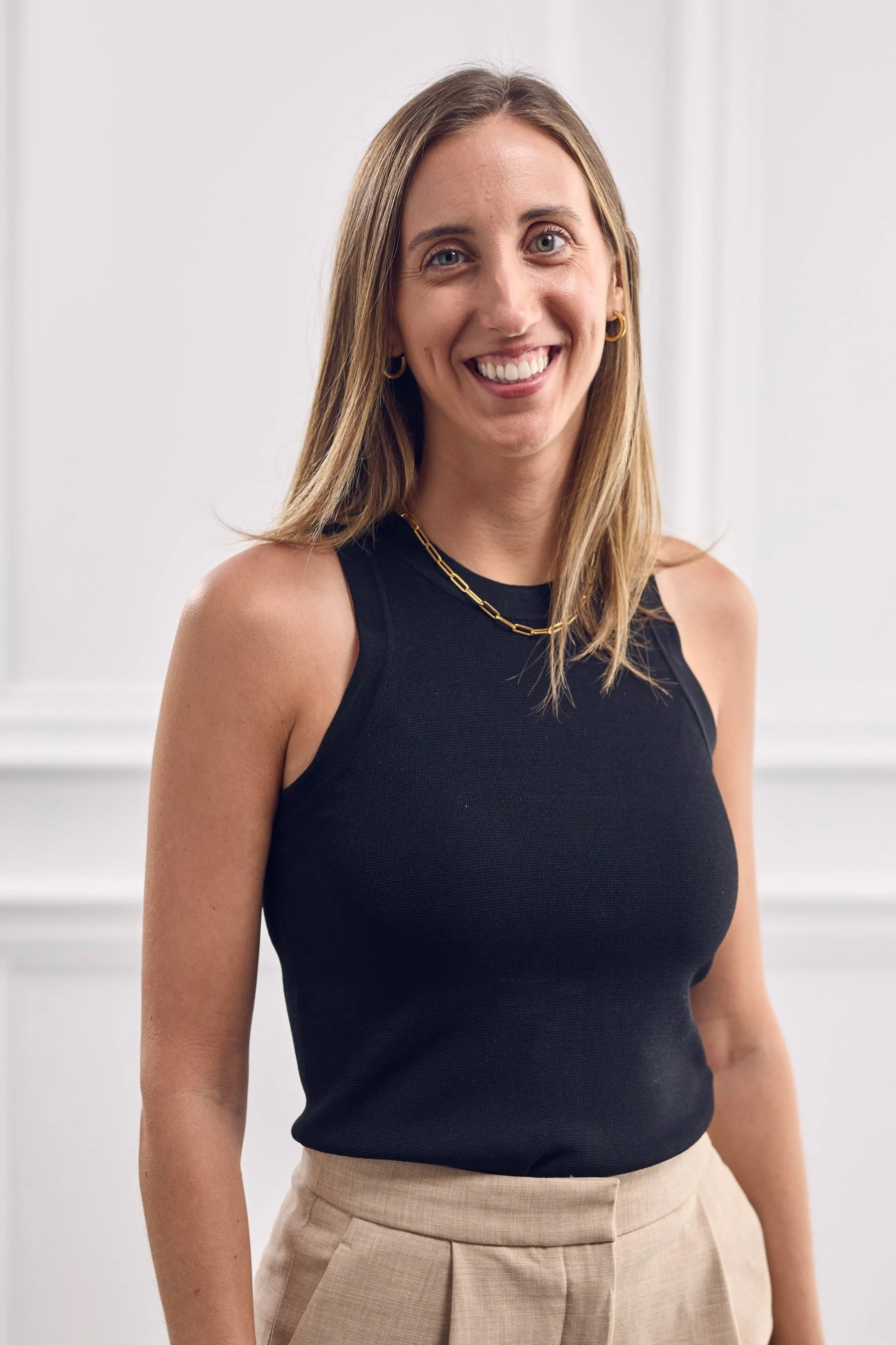 A woman with shoulder-length light brown hair, wearing a black sleeveless top and beige high-waisted pants, standing indoors in front of a white wall.
