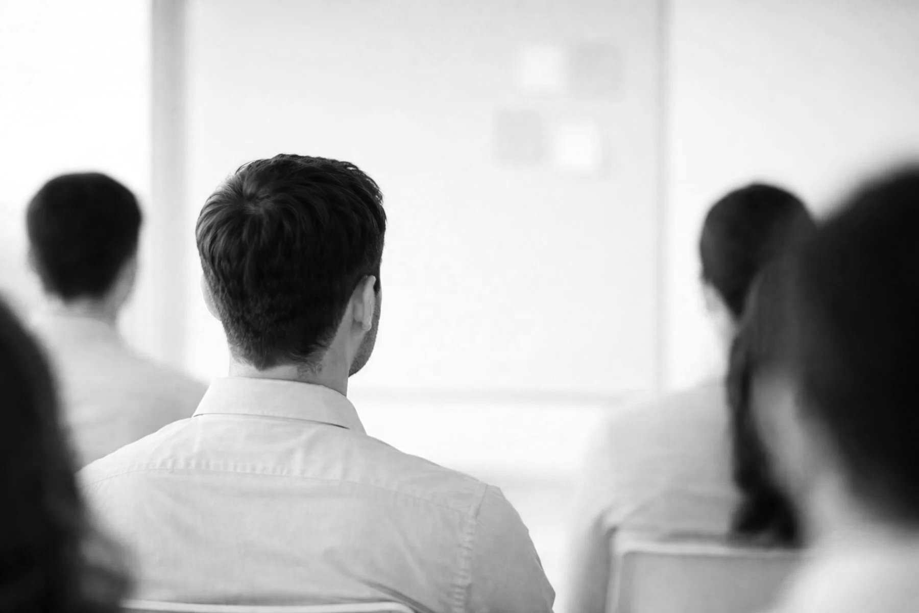 Black and white photo showing the backs of a group of people, focusing on a man with dark hair in the center, seated in a room with a bright background, possibly at a presentation or meeting.
