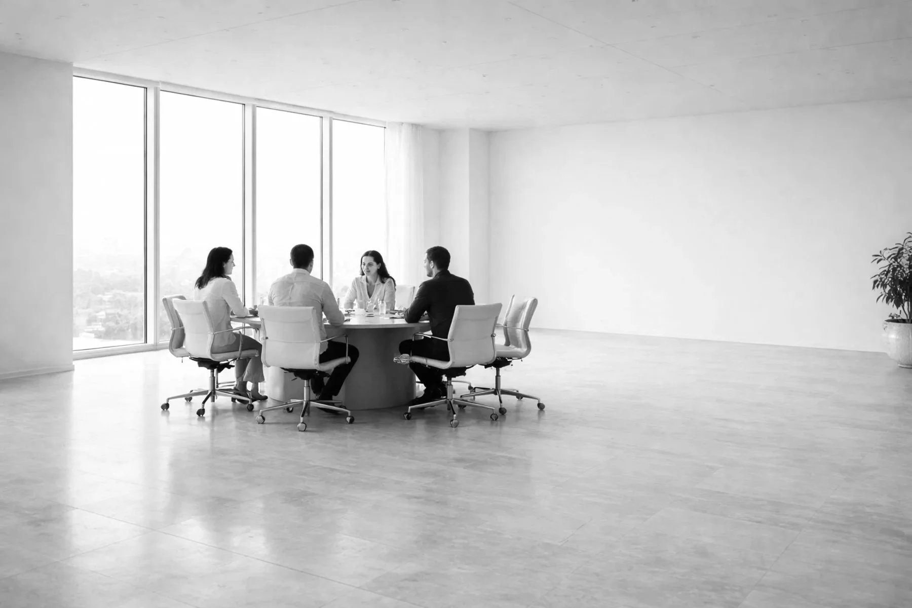 Four people sitting around a round table in a modern, minimalistic conference room with large windows and a potted plant in the corner.