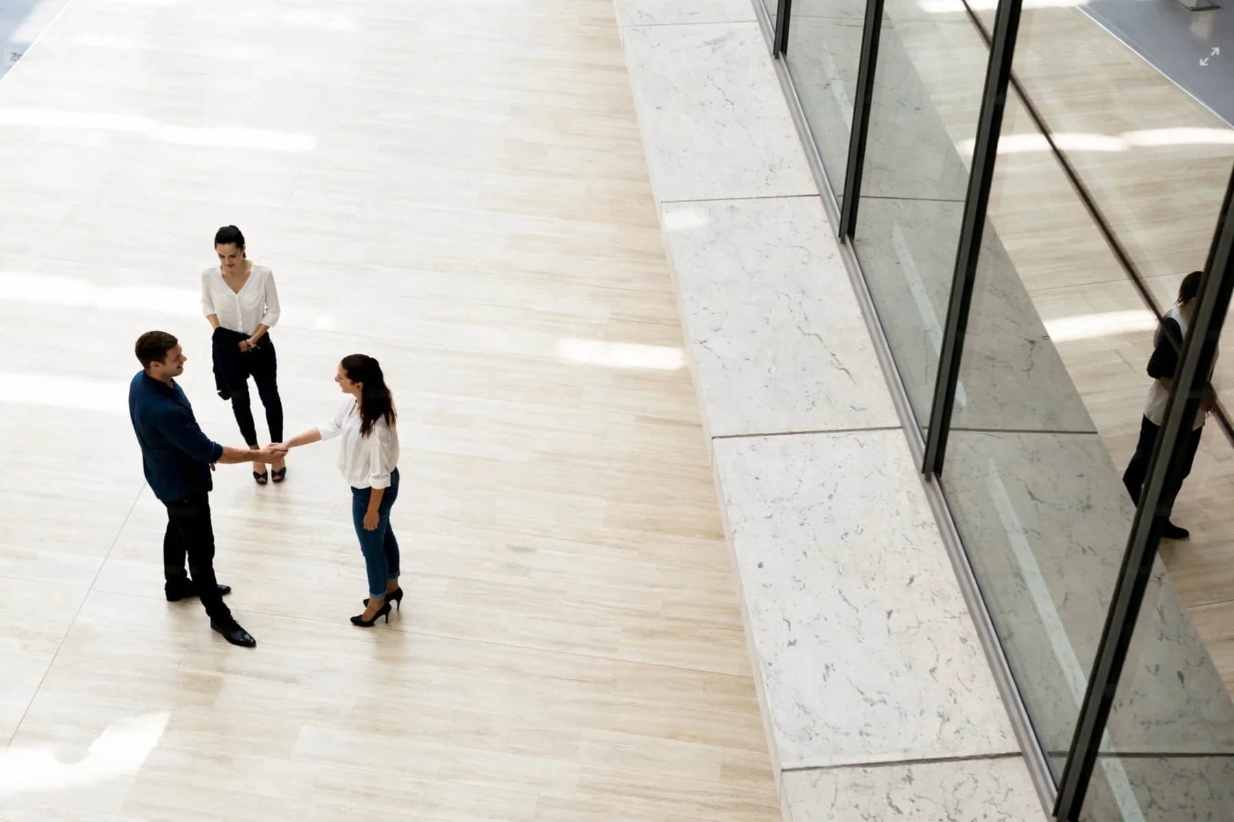 A man and a woman are shaking hands in a spacious, modern interior with a light wooden floor, while a woman in a white blouse and black heels stands nearby. Reflections of the scene can be seen on the glass panels on the right.
