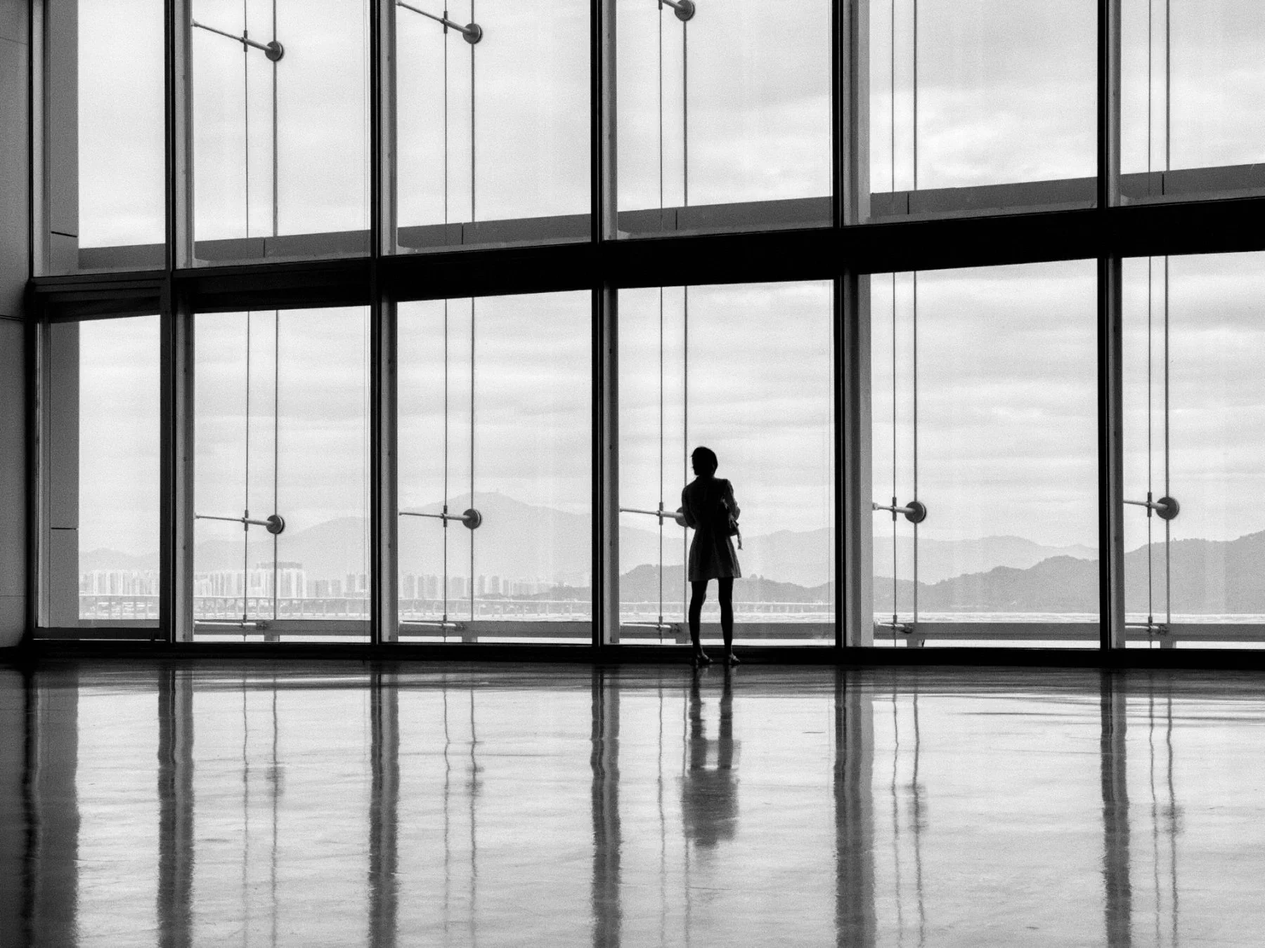 A woman standing inside a modern building with large glass windows, looking outside at a cityscape and mountains in the distance.