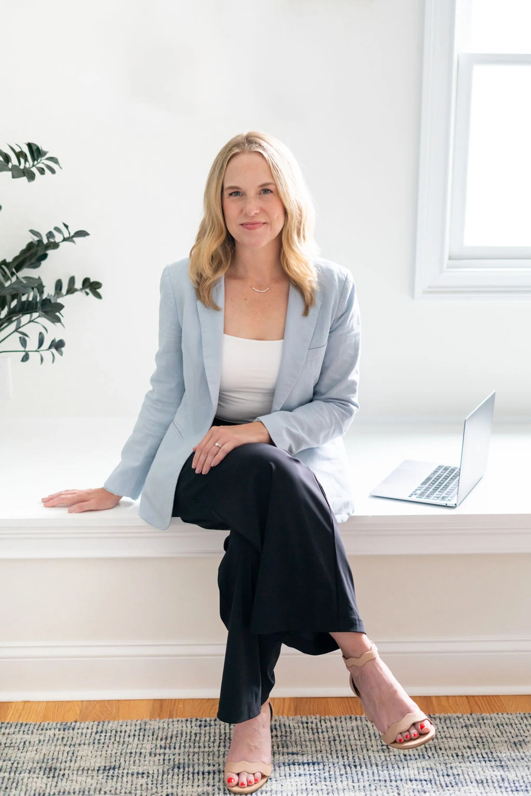 A woman with blonde hair sitting on a white windowsill, wearing a light blazer, black pants, and nude high-heeled sandals, with a laptop on the window ledge and a green plant nearby.