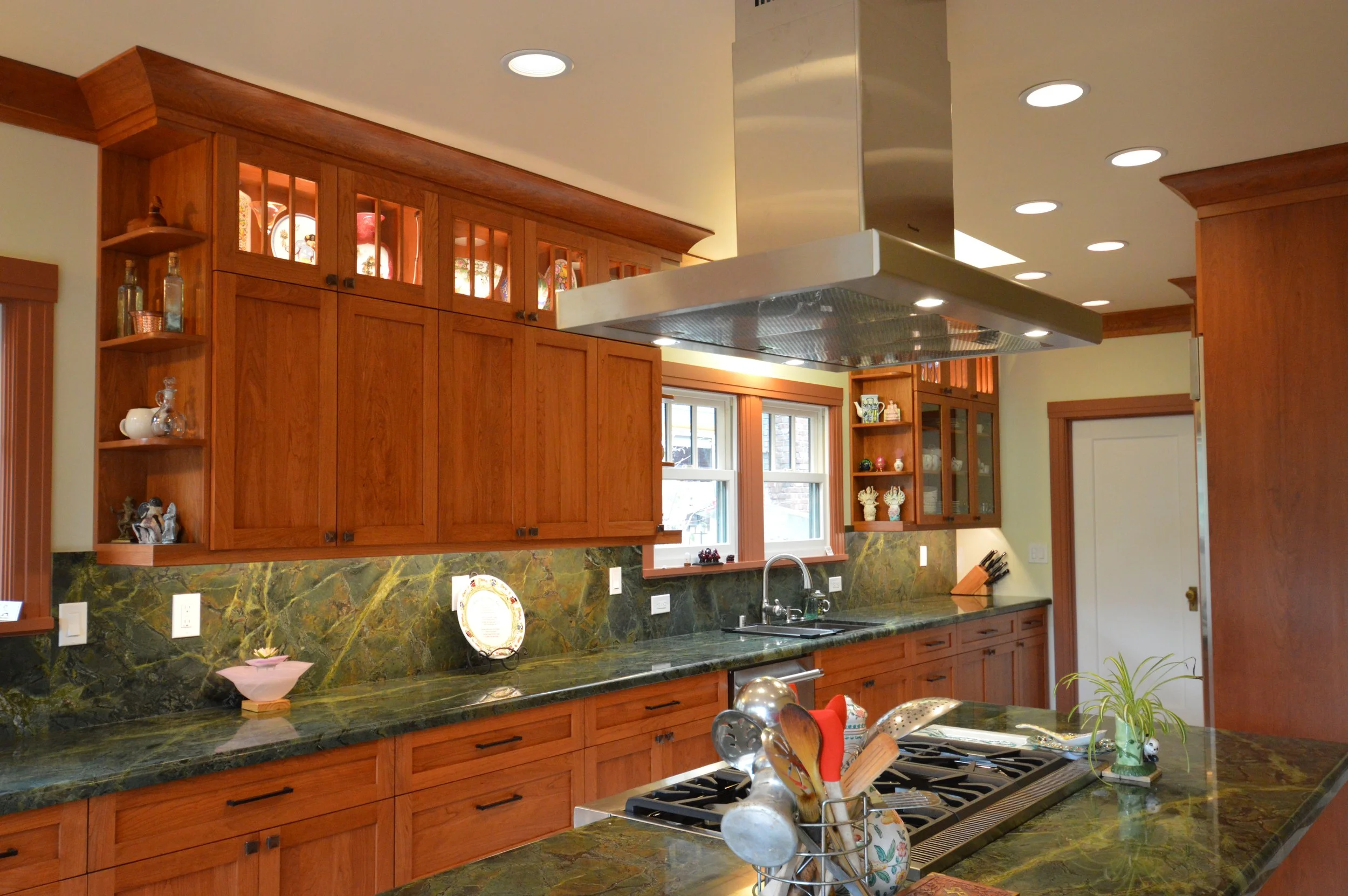 Kitchen with wooden cabinets, green marble countertops, a window above the sink, and a stainless steel range hood. Various decorative items and kitchen utensils are on the counters and shelves.