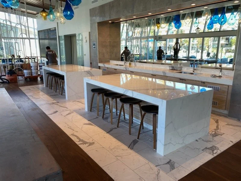 Modern restaurant or cafe interior with marble tables, bar stools, colorful hanging pendant lights, large mirror, and windows with a view outside.