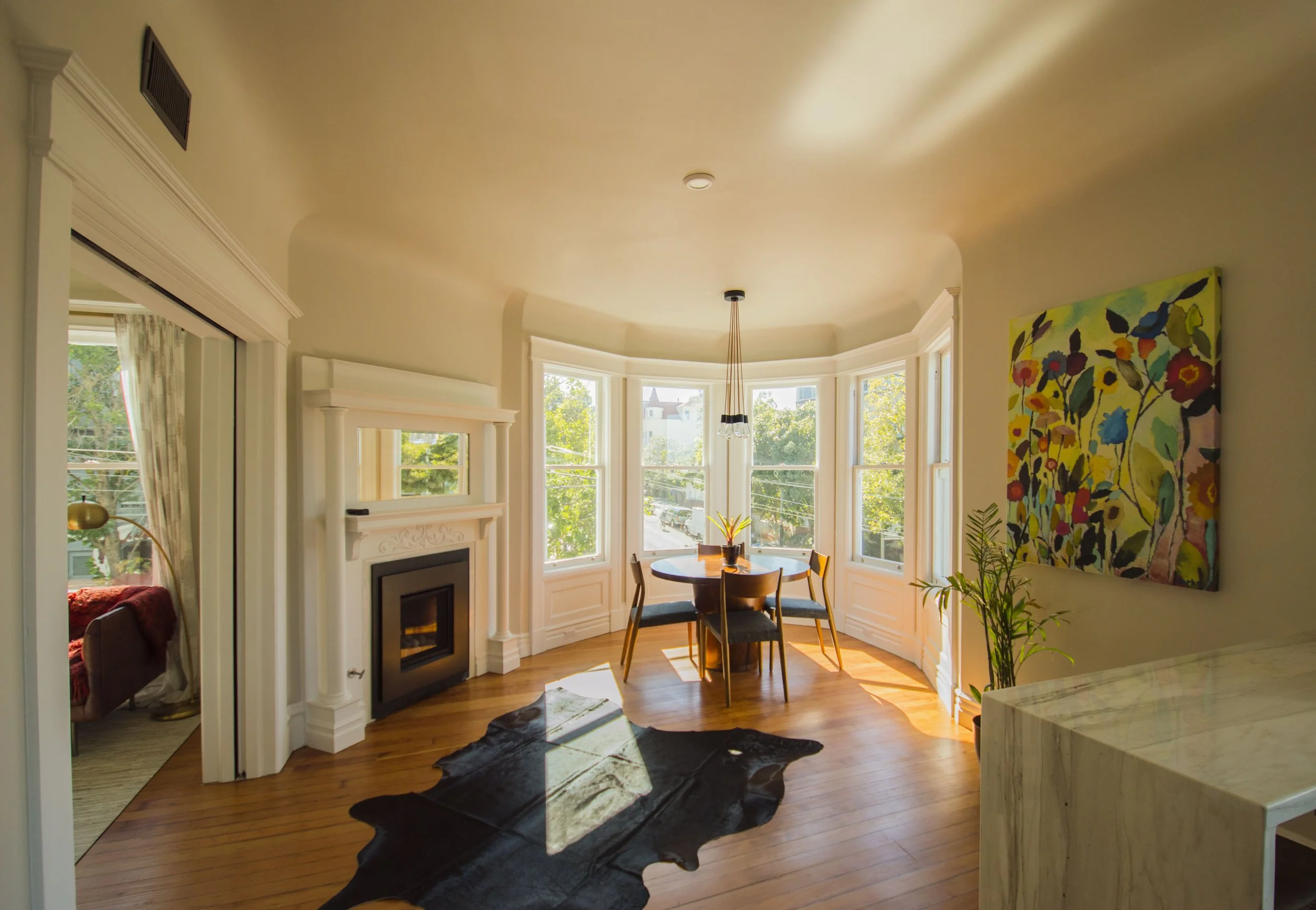 Bright living room with large bay window, white fireplace mantel, colorful floral painting on the wall, and hardwood floors with a black cowhide rug.