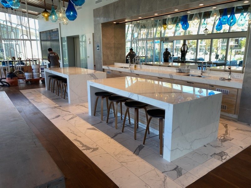 Modern restaurant interior with marble countertops, high stools, hanging glass pendant lights, and large windows.
