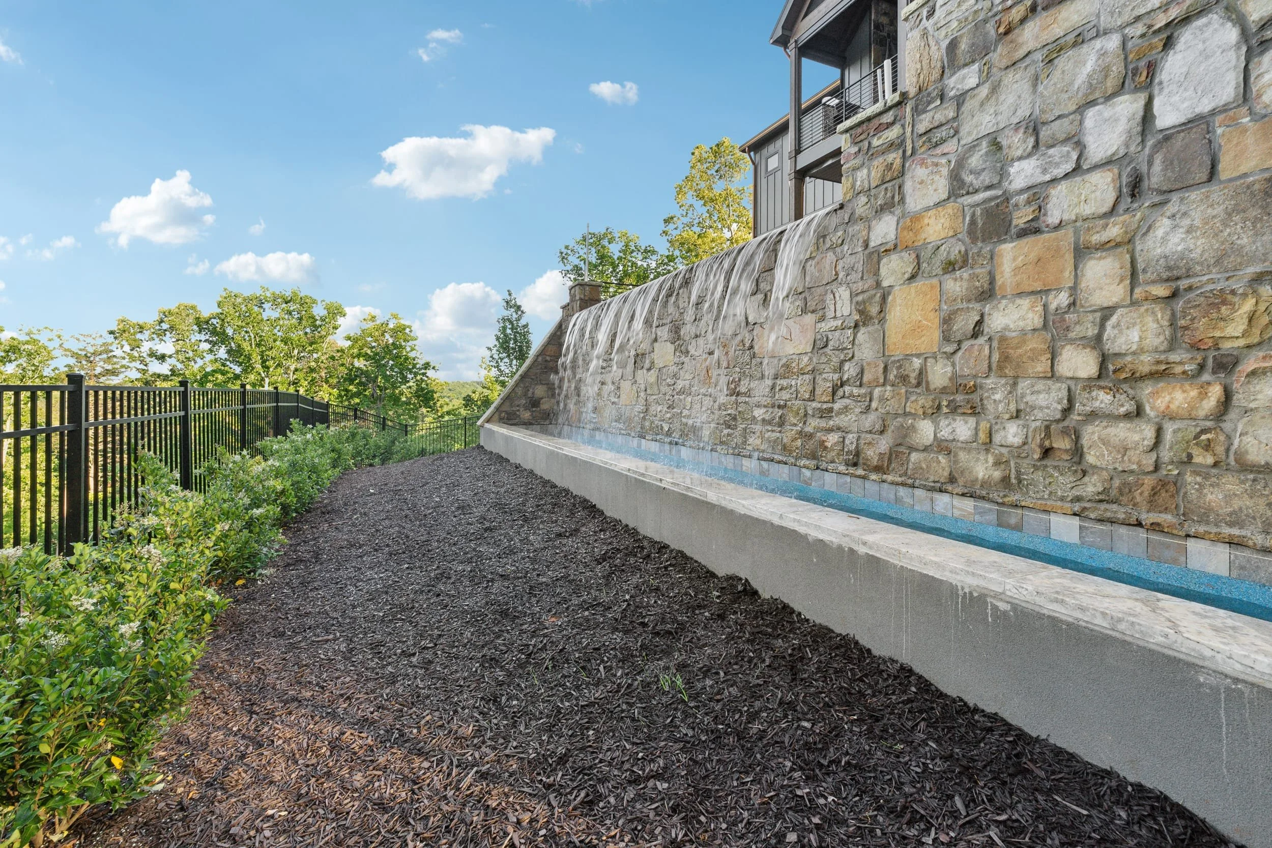 A backyard with a stone retaining wall, a waterfall feature, black metal fence, green bushes, and trees under a blue sky with clouds.