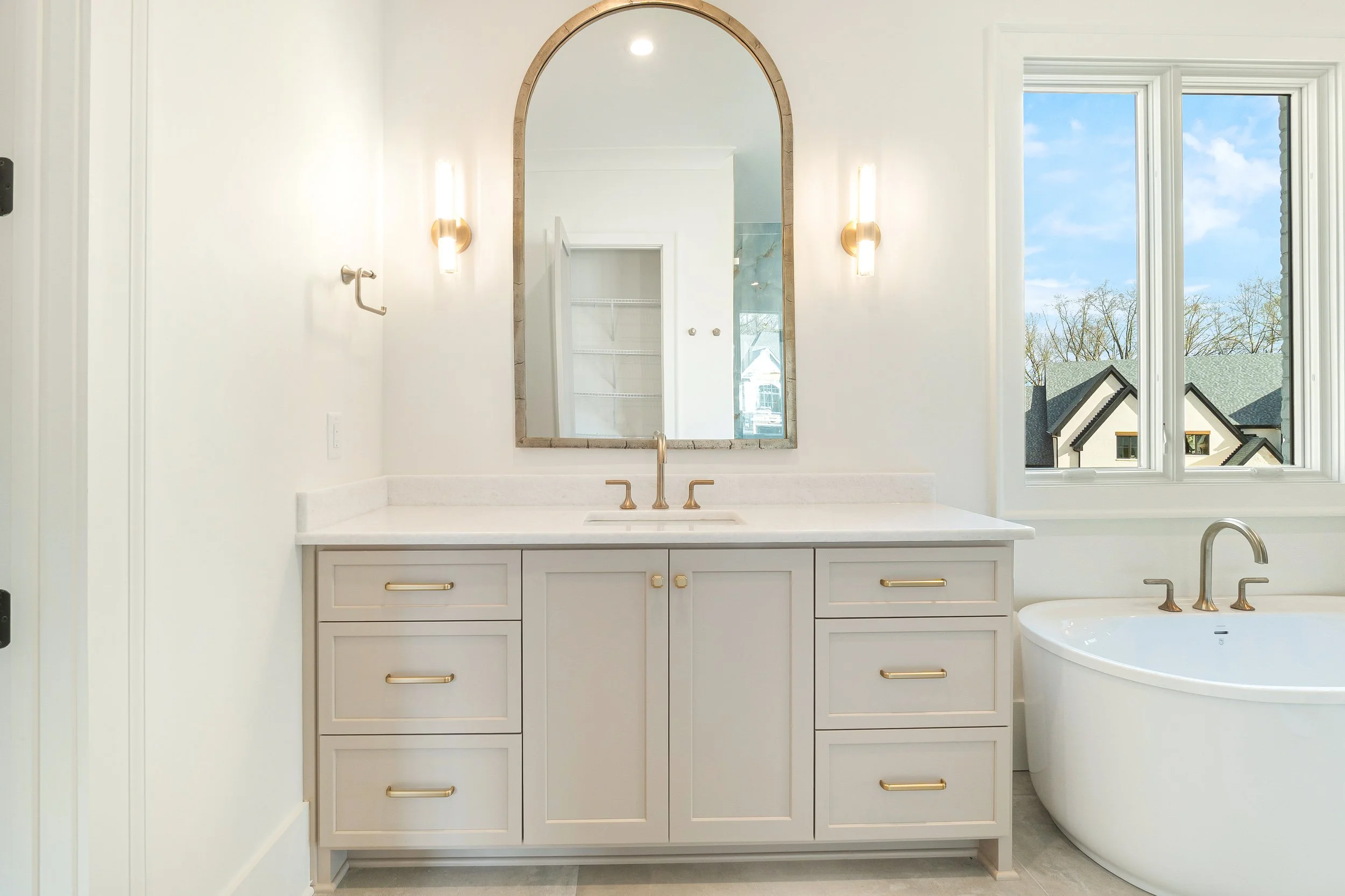 A modern bathroom with a marble vanity, gold fixtures, a large arched mirror, twin vanity lights, and a window showing a suburban neighborhood with trees and houses.
