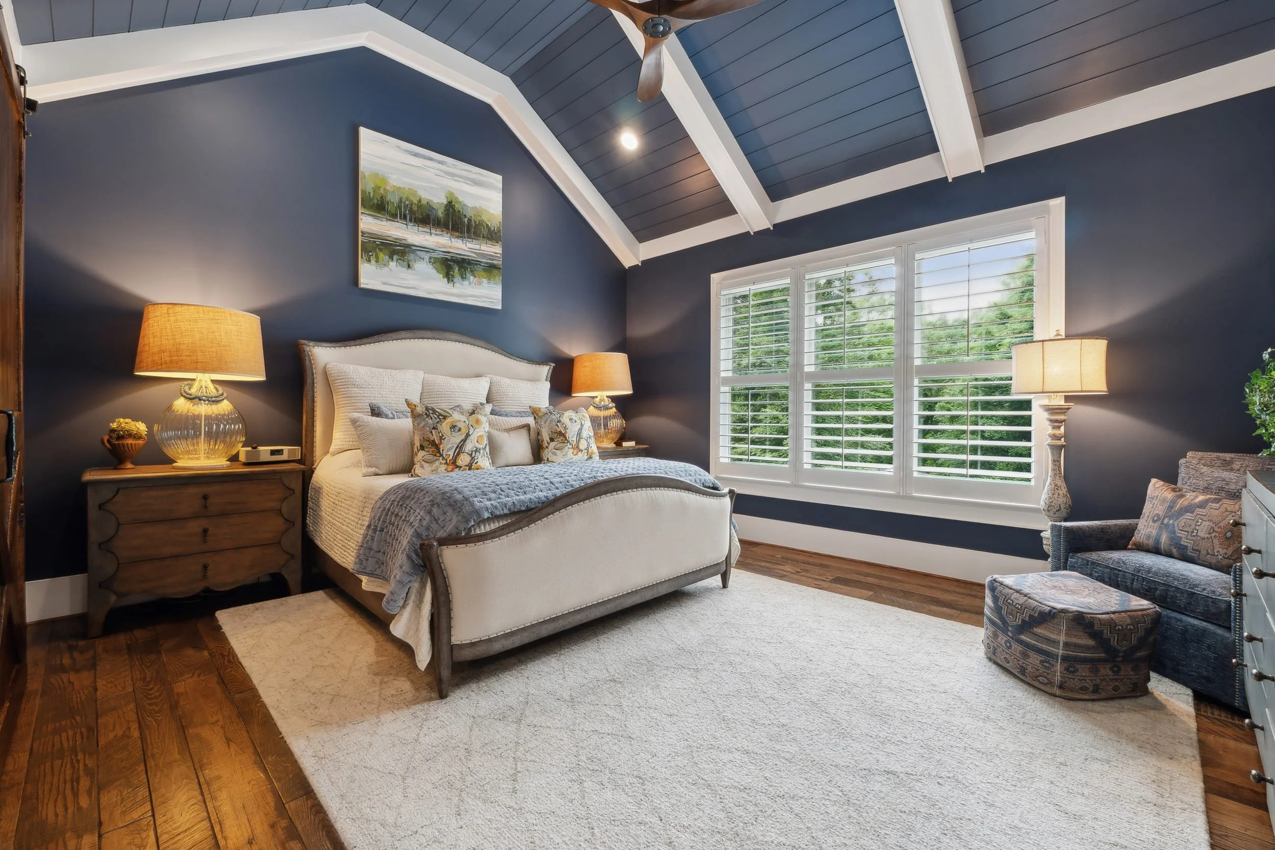 A bedroom with a white upholstered bed, blue walls, and large windows with white shutters. There are bedside tables with lamps, a painting above the bed, a beige area rug, and a patterned armchair.
