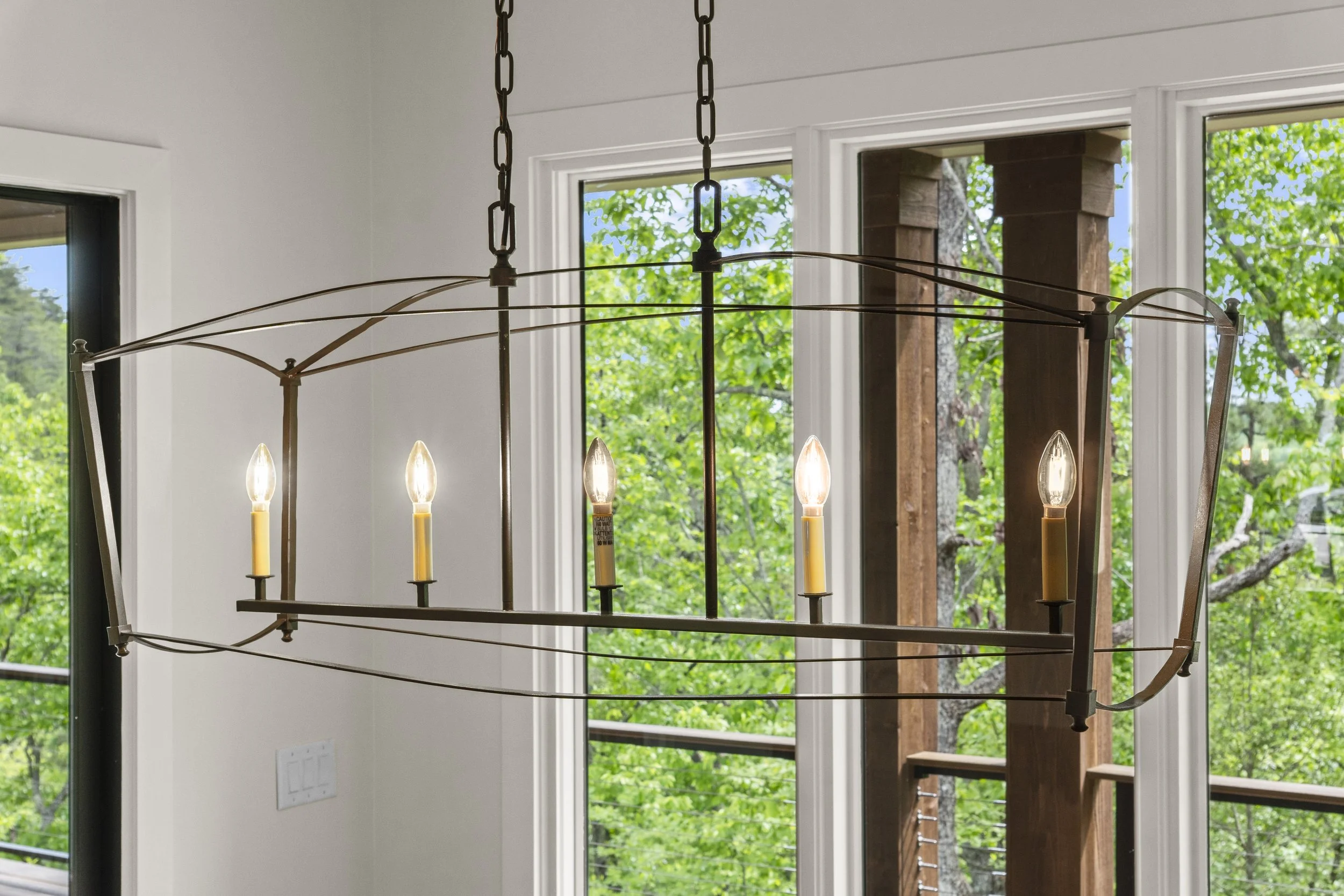 A chandelier with six candle-shaped lights hangs from a chain on a ceiling near large windows showing green trees outside.