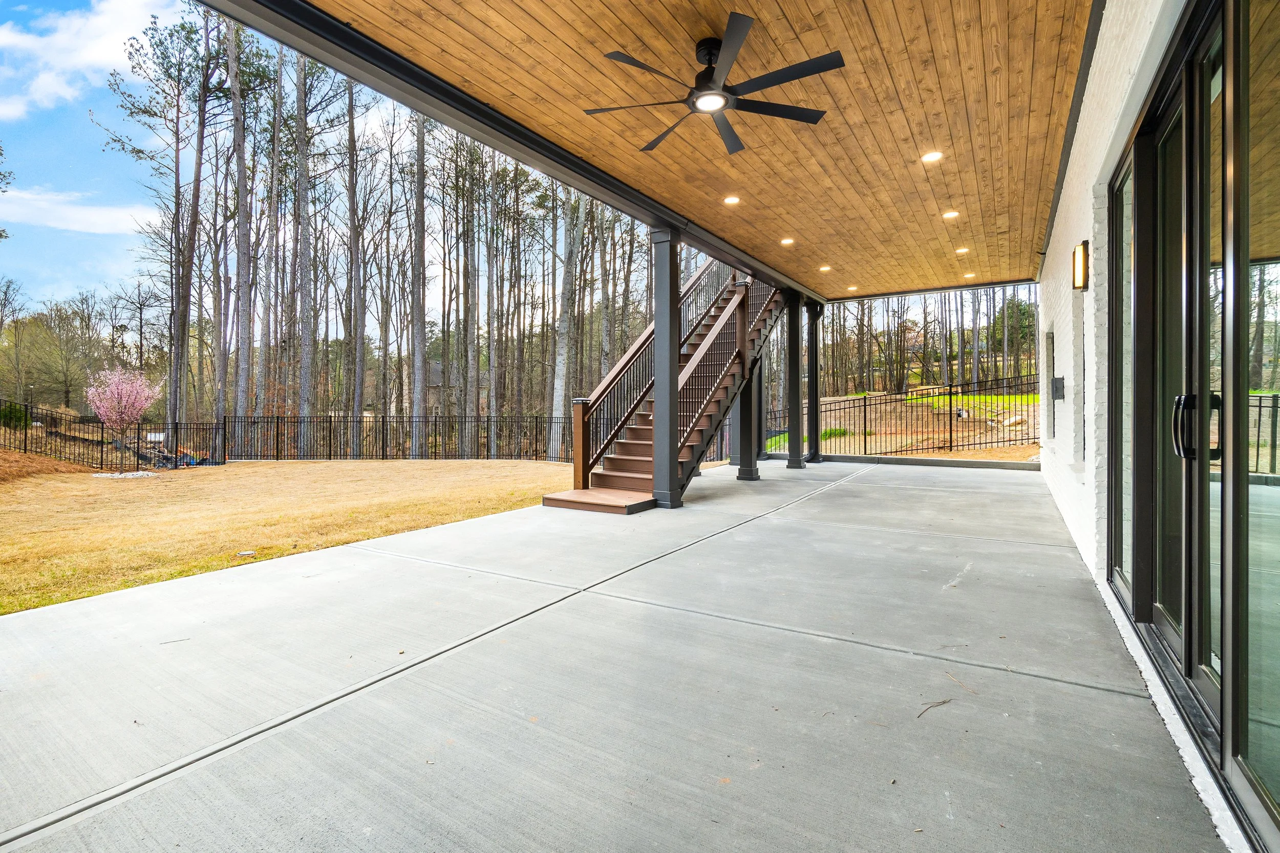 A covered patio area with a concrete floor, wooden ceiling with embedded lighting, ceiling fan, and stairs leading up to the yard. The yard has a tree with pink blossoms and is enclosed by a black metal fence, surrounded by trees and a partly cloudy 