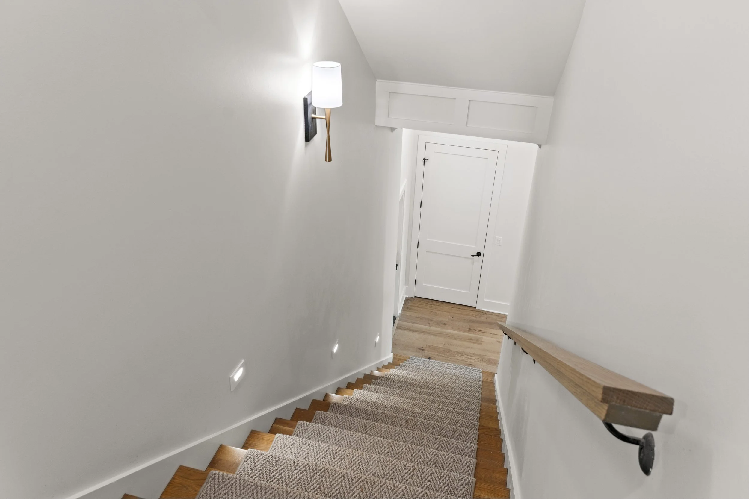 Interior view of a stairway with beige carpet, white walls, and a wooden handrail on the right side. Modern wall-mounted light fixtures illuminate the hallway, leading to a closed white door at the top of the stairs.