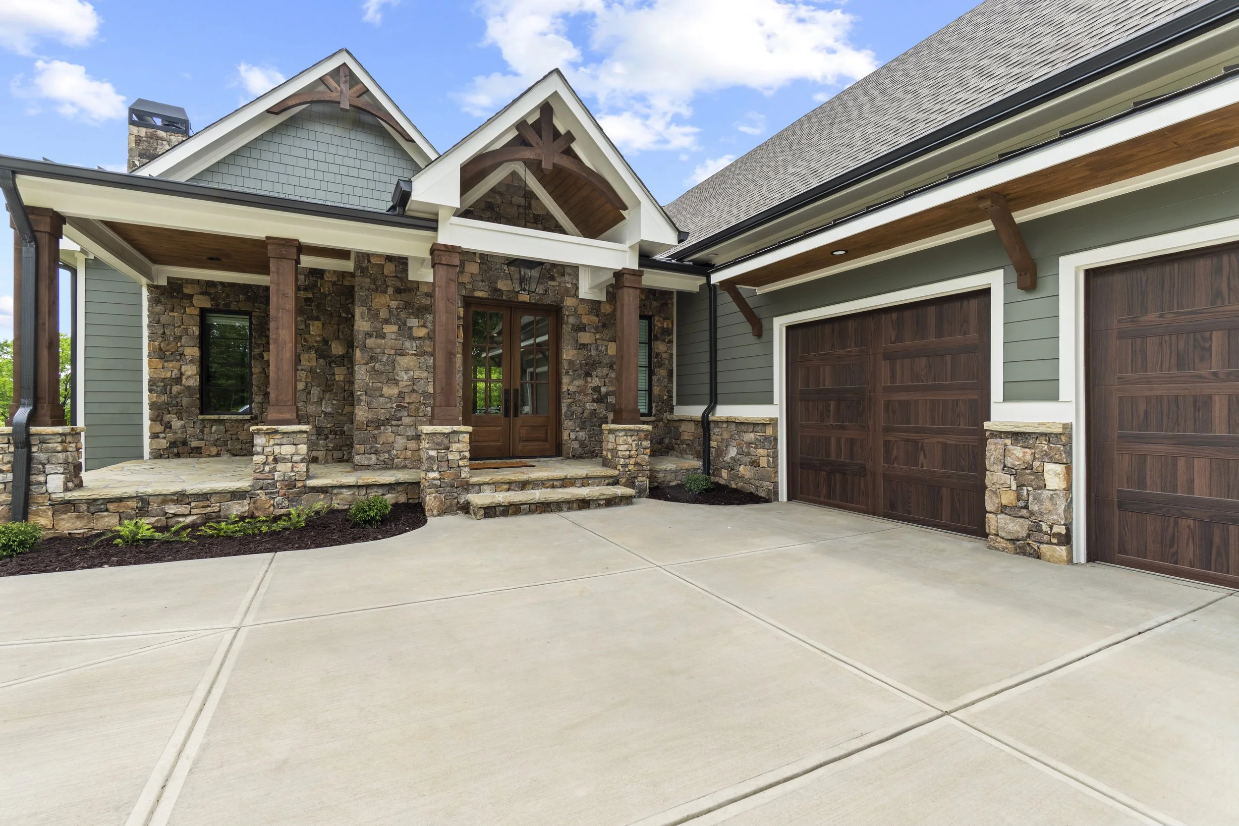 Front view of a house with stone and siding exterior, double garage doors, stone steps, and a concrete driveway, clear sky with clouds
