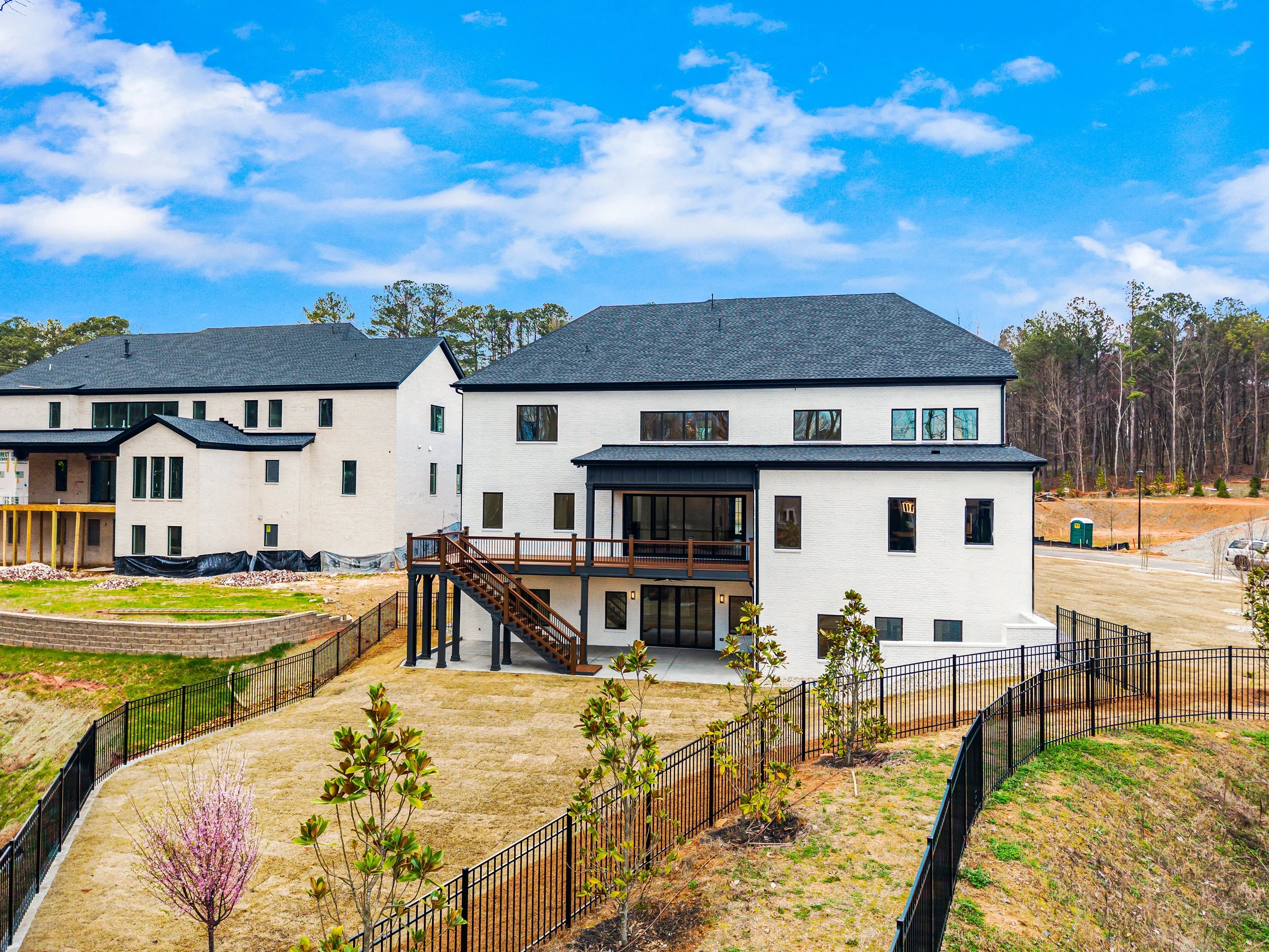 Newly constructed residential houses with large windows, a wooden staircase, and fenced yards under a partly cloudy blue sky.