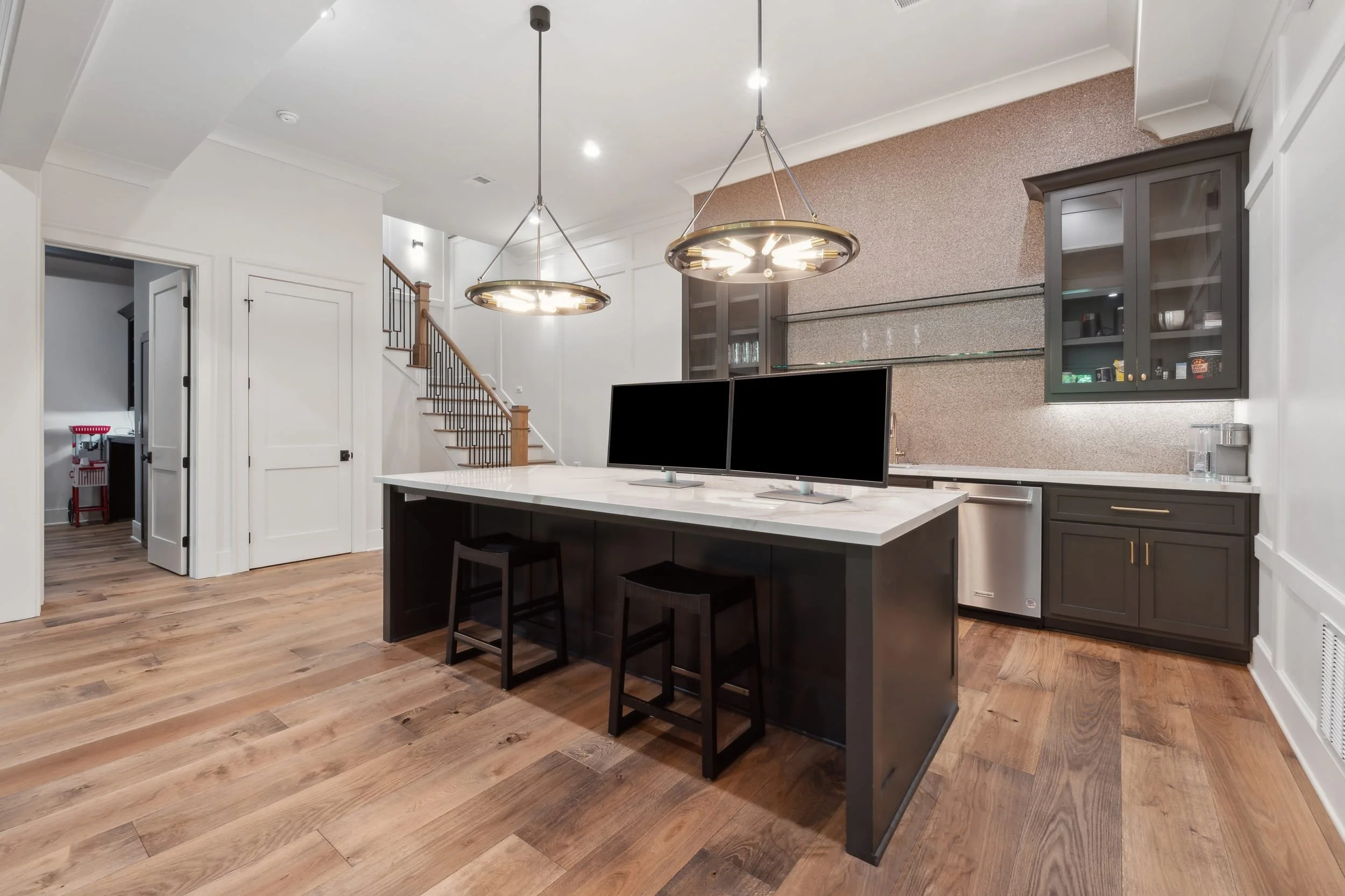 Modern kitchen with black island, dual computer monitors, pendant lights, gray cabinets, and wooden flooring.