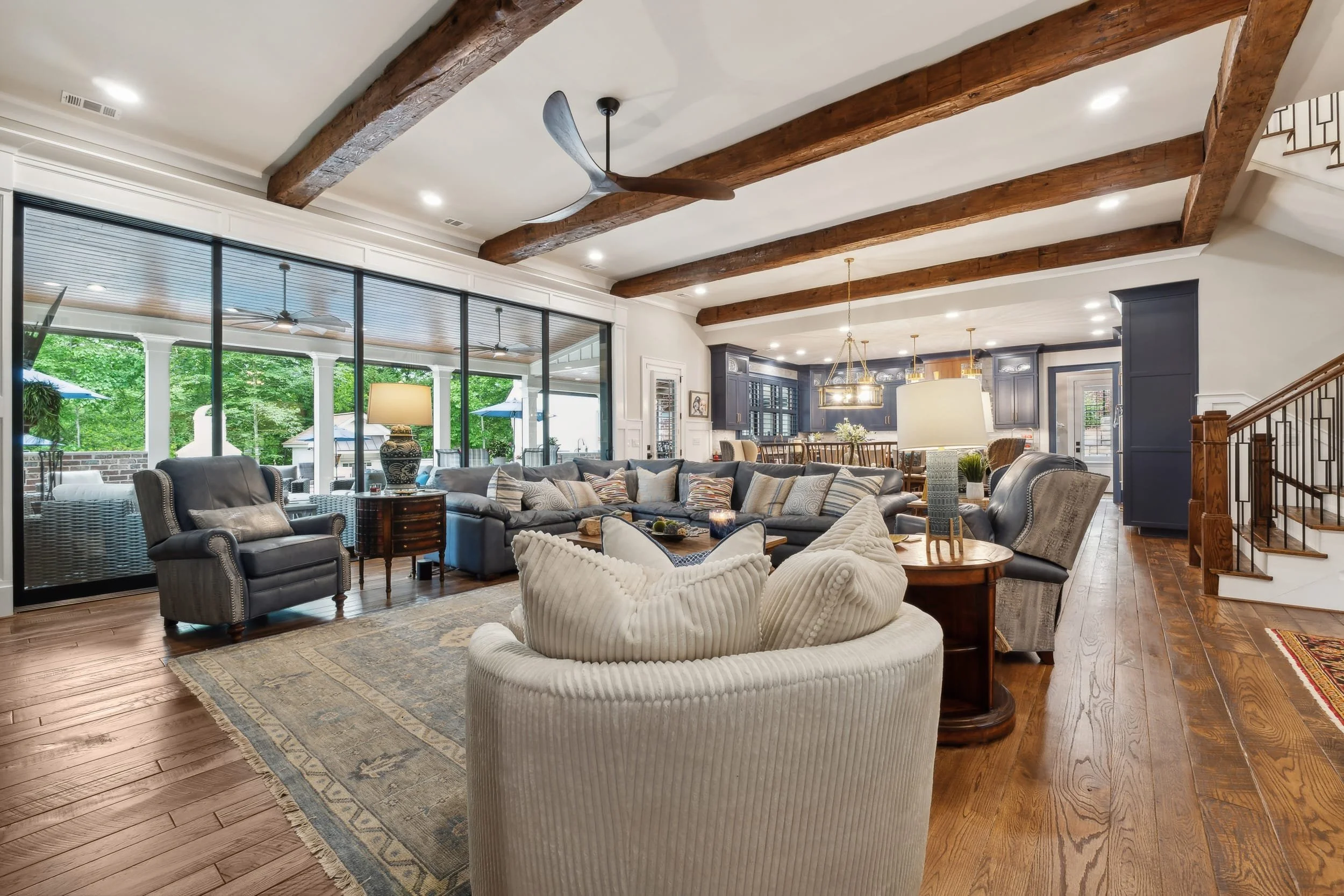 A spacious living room with wood beams on the ceiling, large windows, a gray sectional sofa, and a mix of chairs and tables. The room is open to a dining and kitchen area with dark cabinets and pendant lighting.