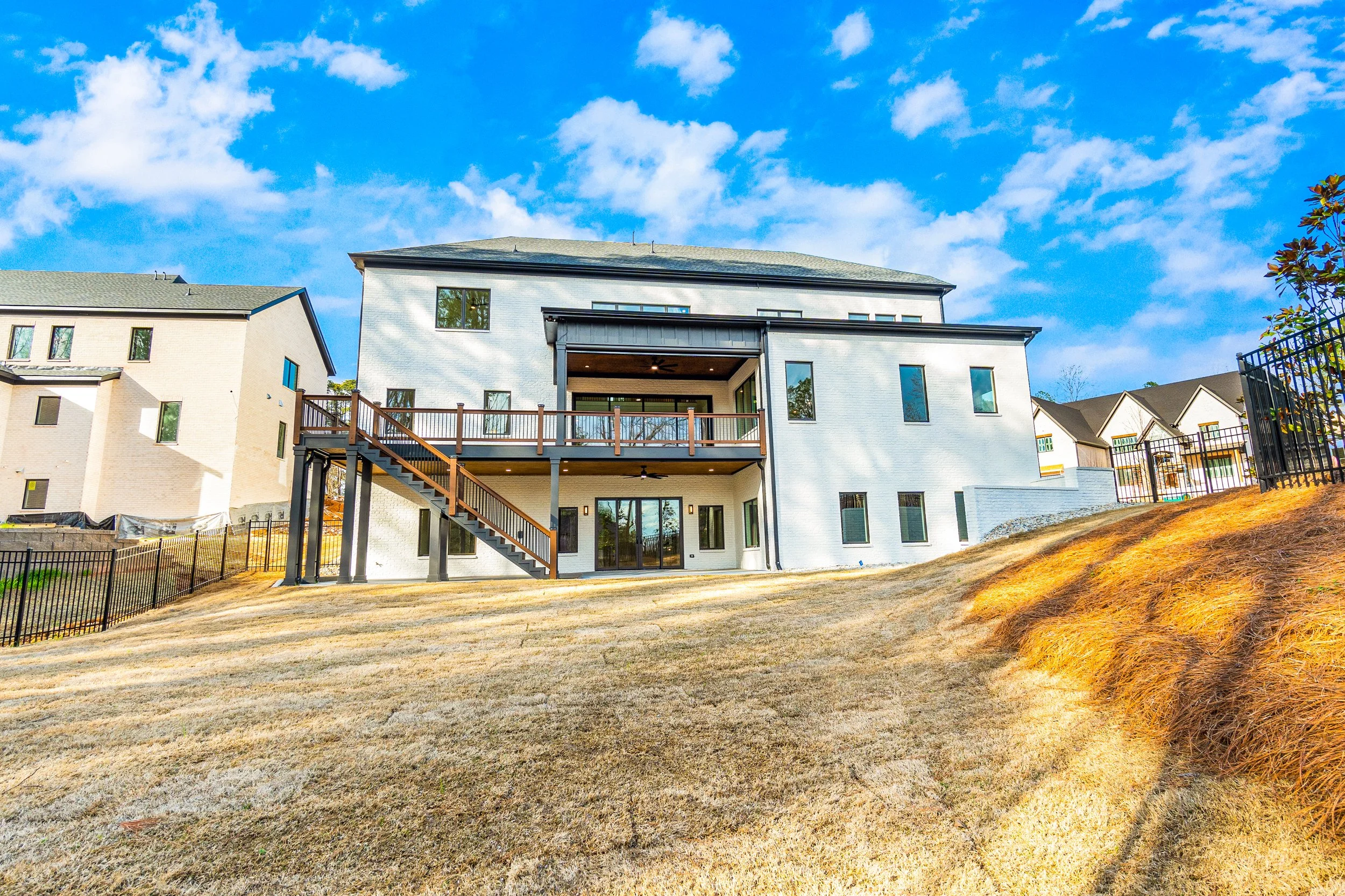 Modern white house with multiple balconies, set on a grassy hill, under a bright blue sky with scattered clouds.