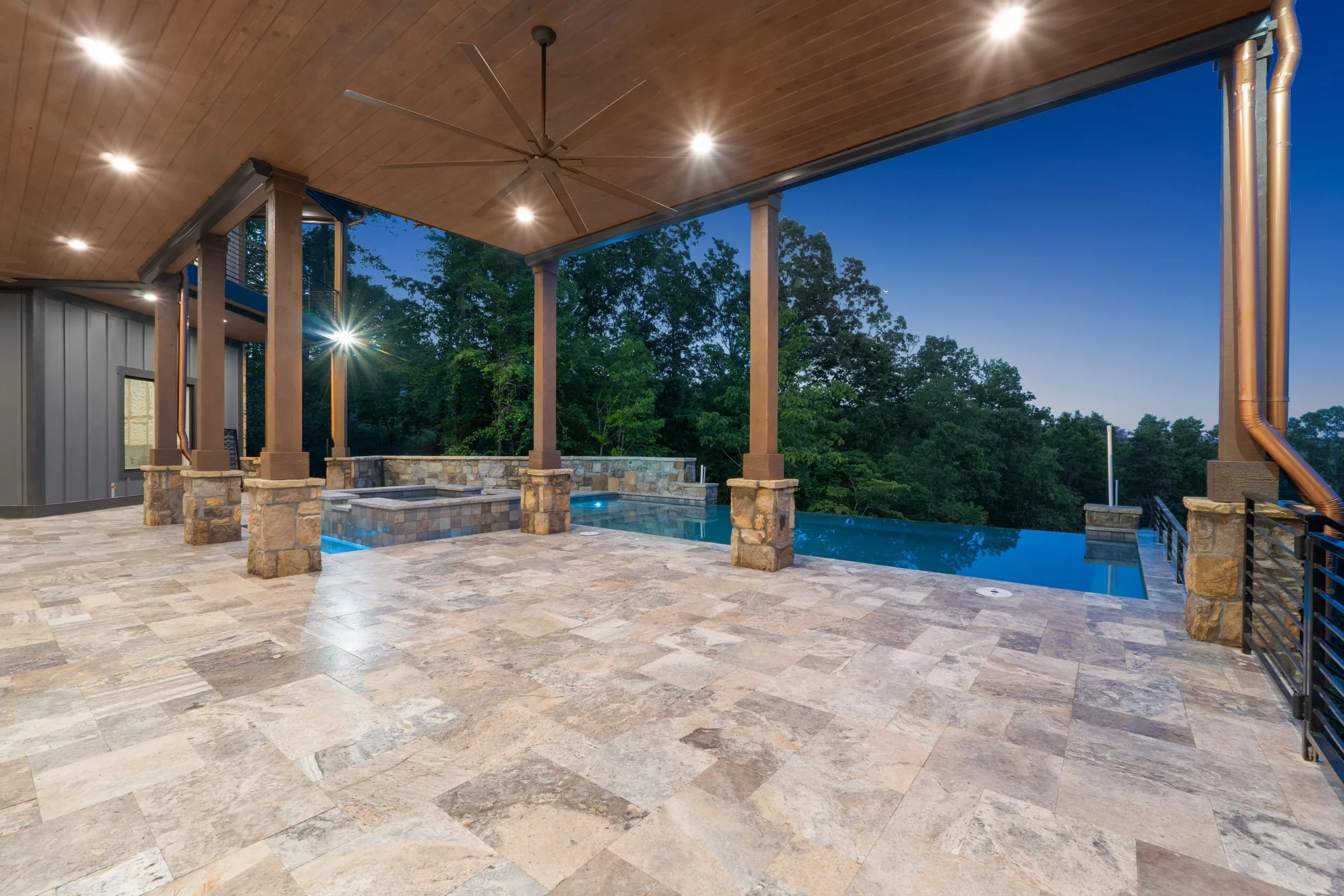  outdoor covered patio with stone flooring, stone pillars, a hot tub, ceiling fan, lighting, and a view of trees and the evening sky in the background