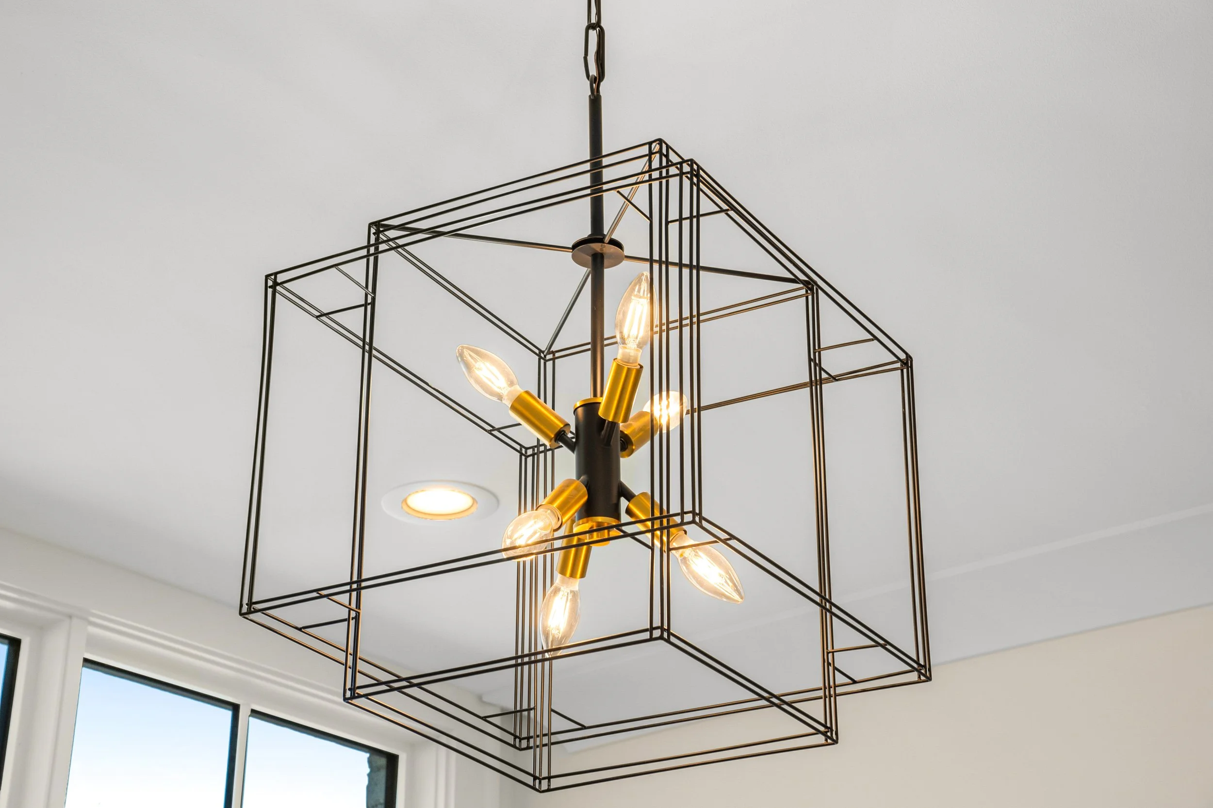 A modern black metal chandelier with exposed light bulbs hanging from a chain against a white ceiling and adjacent window.