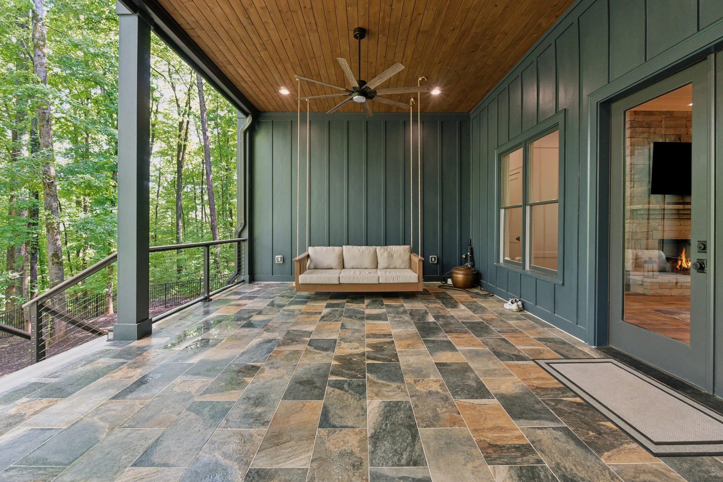 A covered outdoor porch with slate tile flooring, a beige swing, a bronze pot, ceiling fan, and windows overlooking a lush green forest.