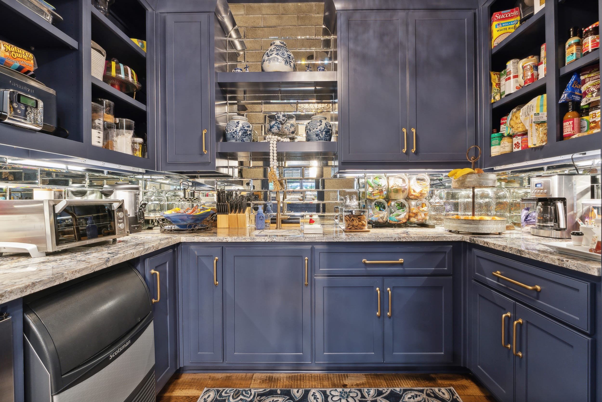 A kitchen with blue cabinets, a granite countertop, and open shelves containing various jars, bottles, and kitchen items. There is a stainless steel oven and microwave, a knife block, and a backsplash made of reflective tiles. Decor items include blu