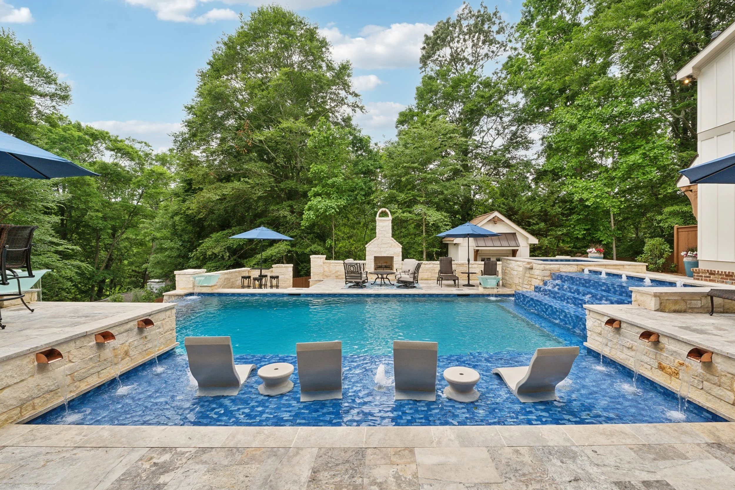 A backyard with an in-ground swimming pool, surrounded by stone patio and chairs, with trees and a blue sky in the background.