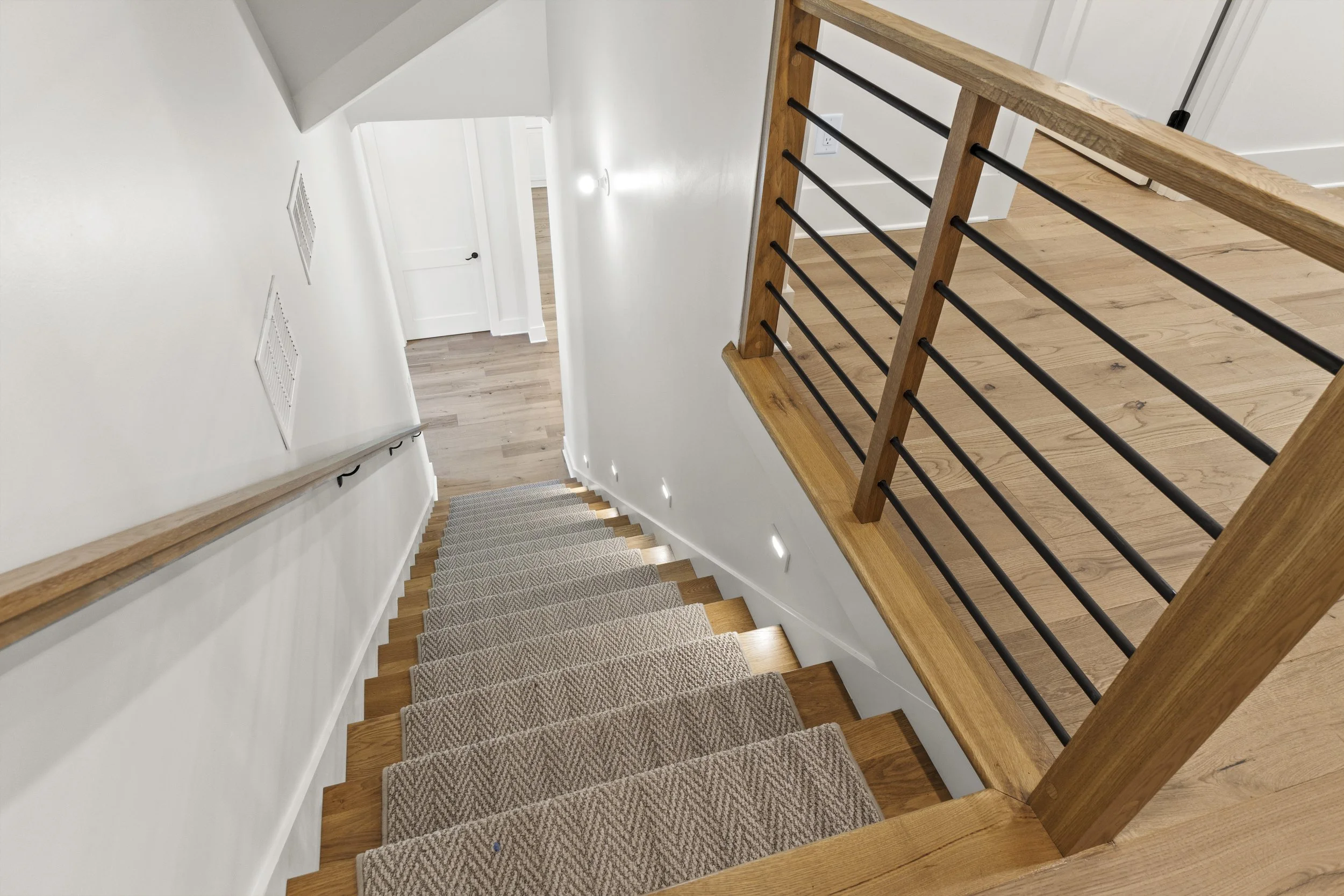 view of a staircase from the top, showing wooden steps with a grey and white patterned carpet runner, a wooden handrail, black metal balusters, white walls, and a closed white door at the bottom.
