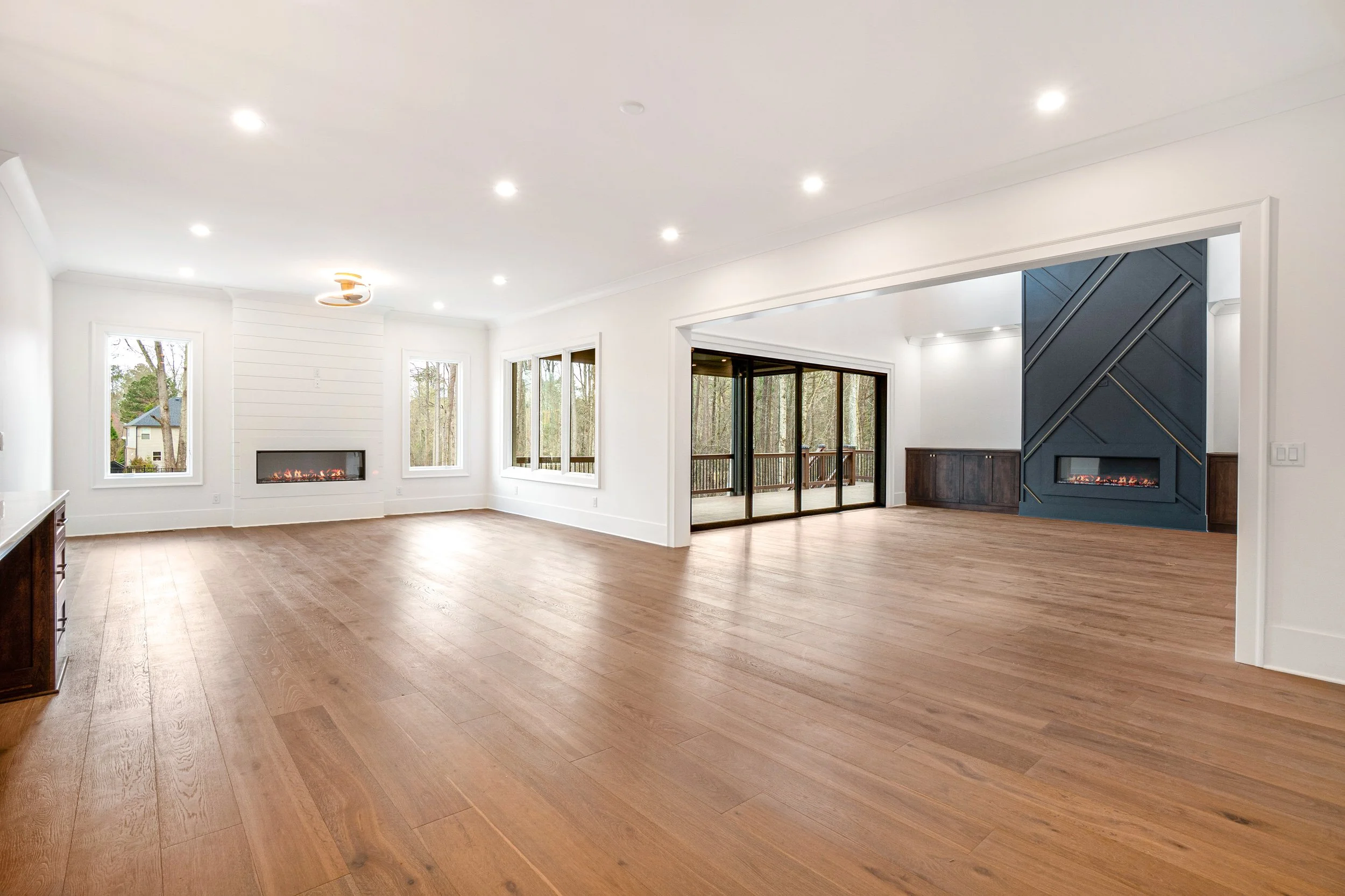 An empty living room with light wood flooring, white walls, multiple windows, a modern fireplace, and a sliding door leading to an outdoor deck with a wooded area beyond.