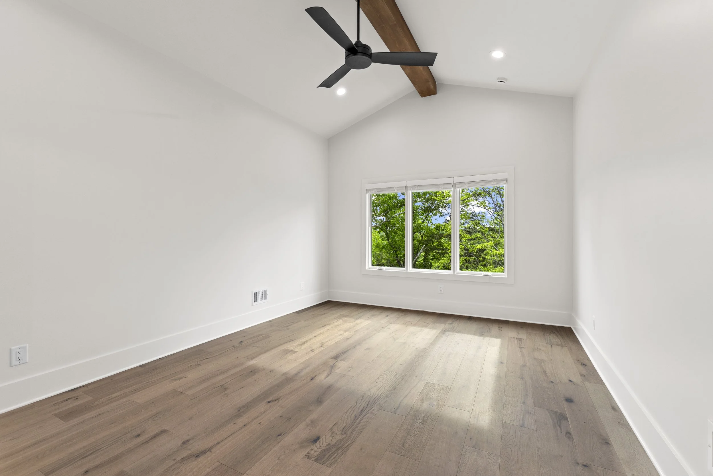 Empty room with white walls, a large window showing green trees outside, hardwood floors, a ceiling fan, and recessed lighting.