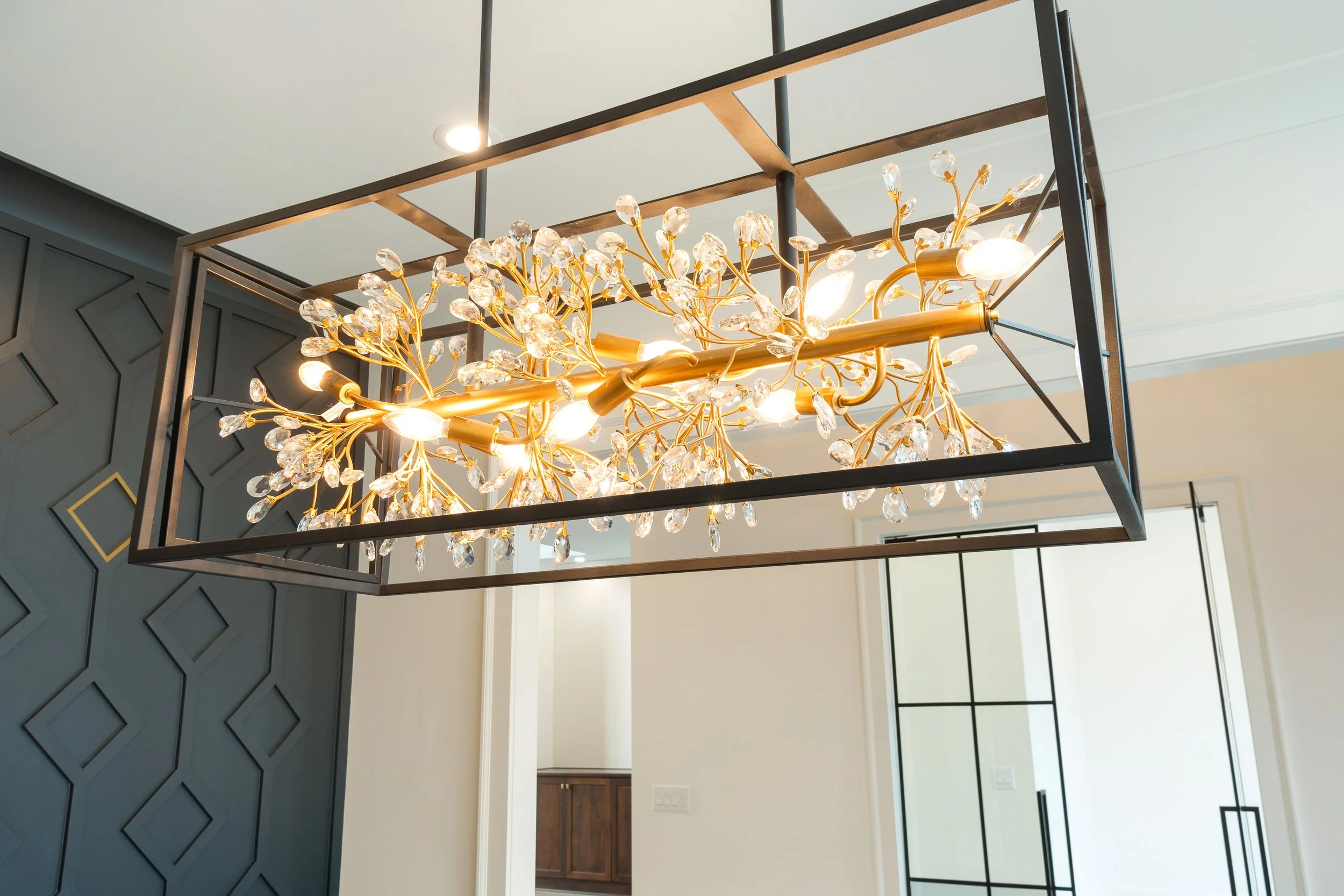 A modern ceiling chandelier with a rectangular frame and decorative gold branches with crystal-like accents, hanging above a room with dark paneled wall and a white ceiling.