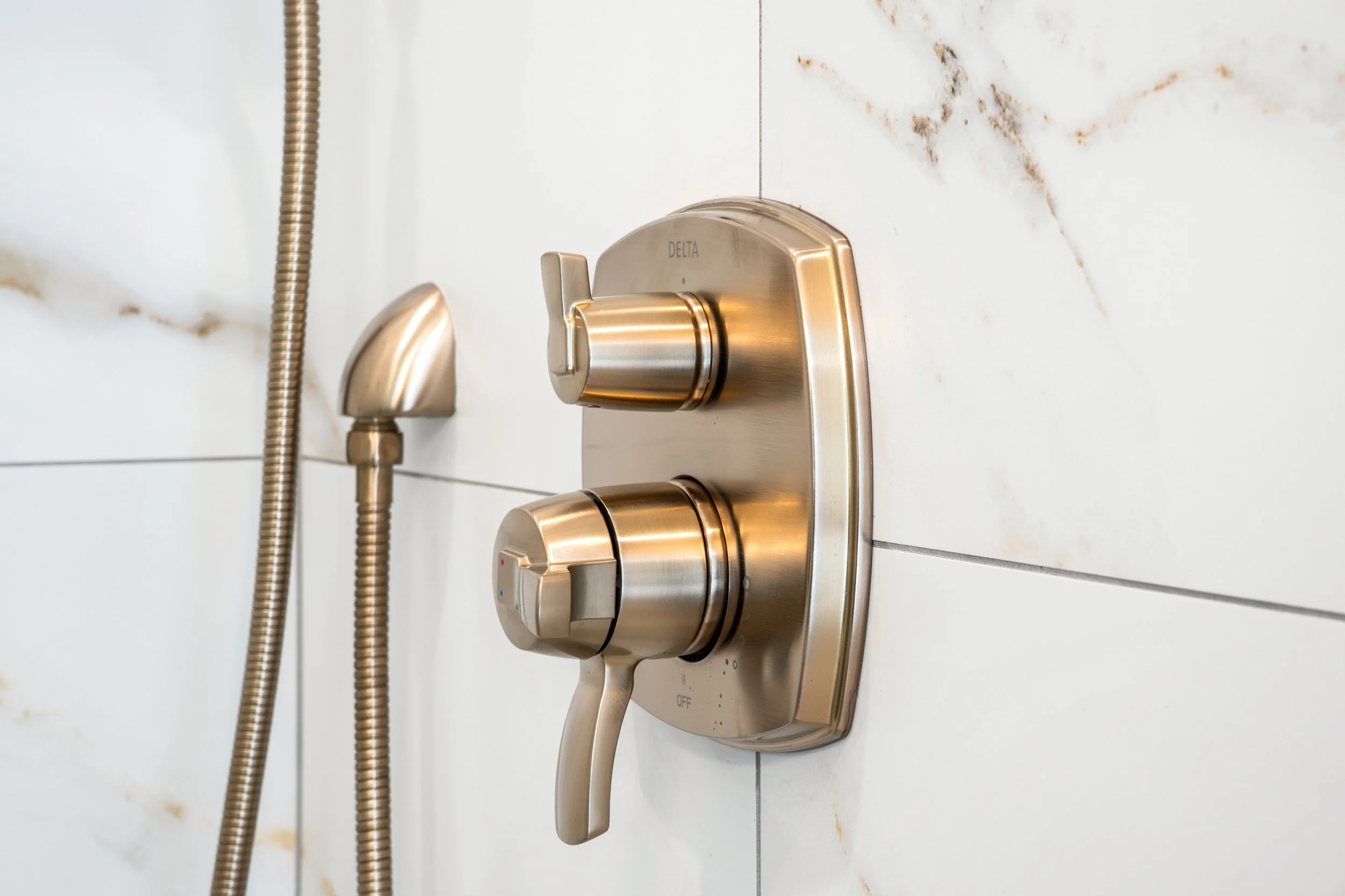 Close-up of a modern, brass-colored shower control panel with a handle and two dials mounted on a white tiled wall with brown veining.