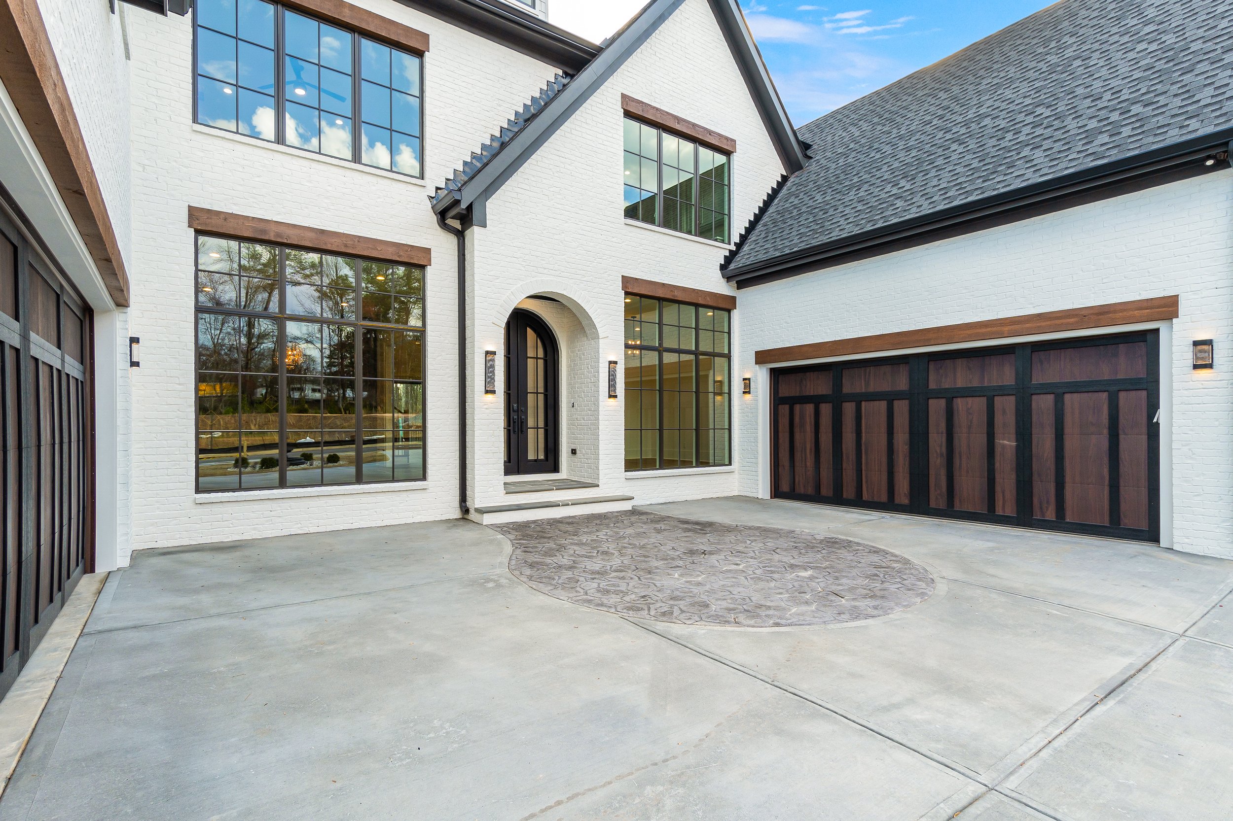 Front view of a modern house with white brick walls, large multi-paned windows reflecting a blue sky with clouds, a black front door with a rounded top, and a attached garage with dark wood and black accents. The driveway is paved with concrete and features a stamped circular pattern.