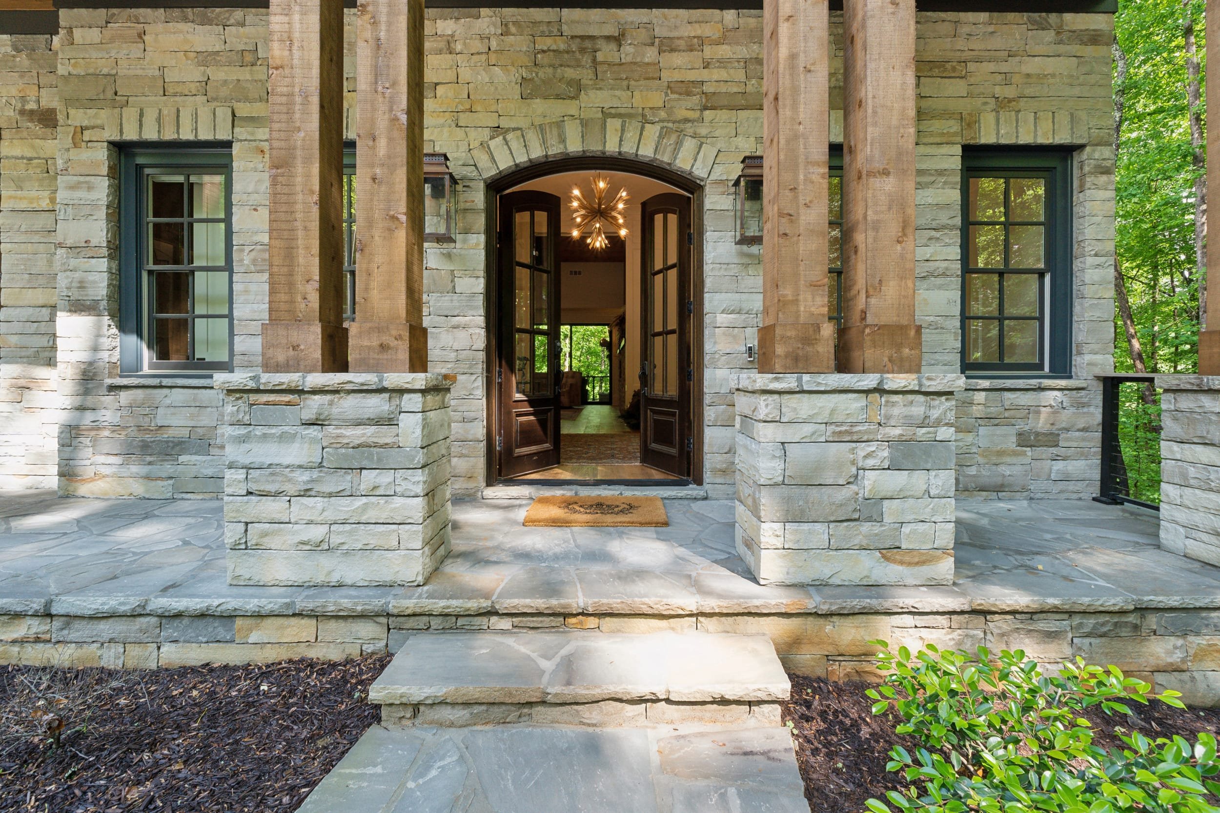 Front porch of a house with stone and wood exterior, open double doors, and two lantern-style wall lights, surrounded by green trees.