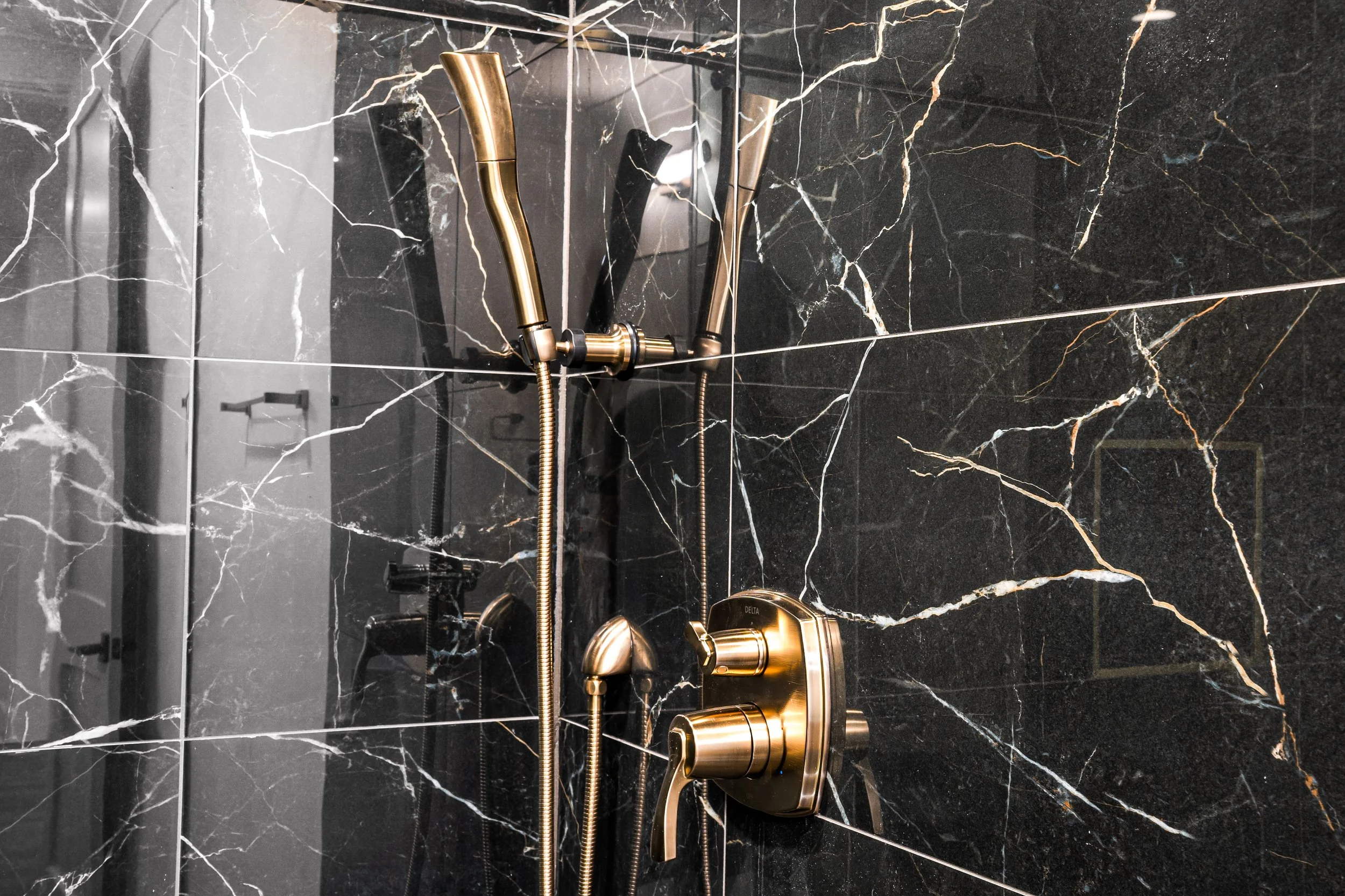 Close-up of a brass shower faucet and handheld showerhead mounted on a black marble wall with white veining.