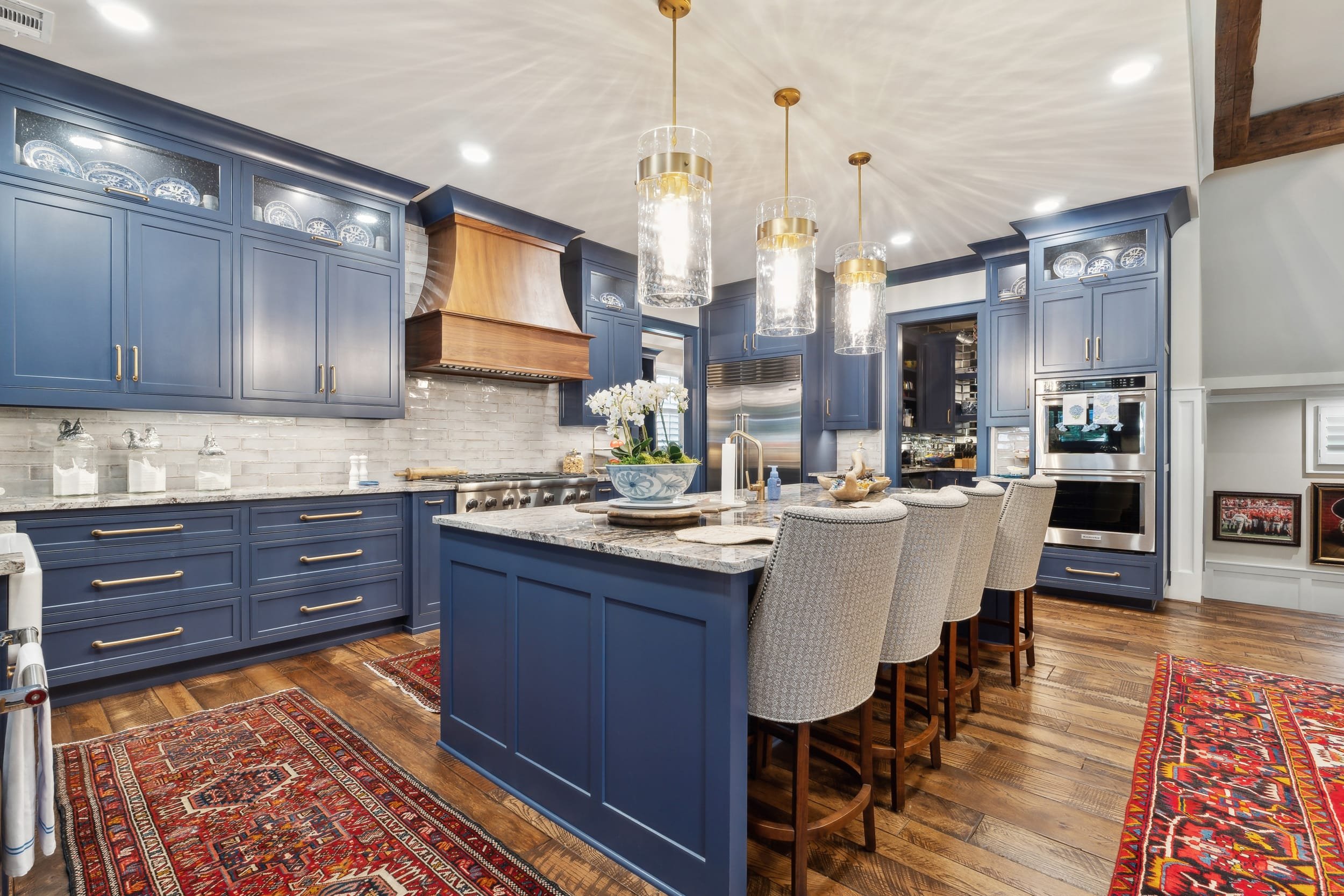 A kitchen with blue cabinets, a marble countertop island, three hanging glass and gold lights, a white brick backsplash, a stainless steel oven, a vase with white flowers, and patterned area rugs on wooden floors.