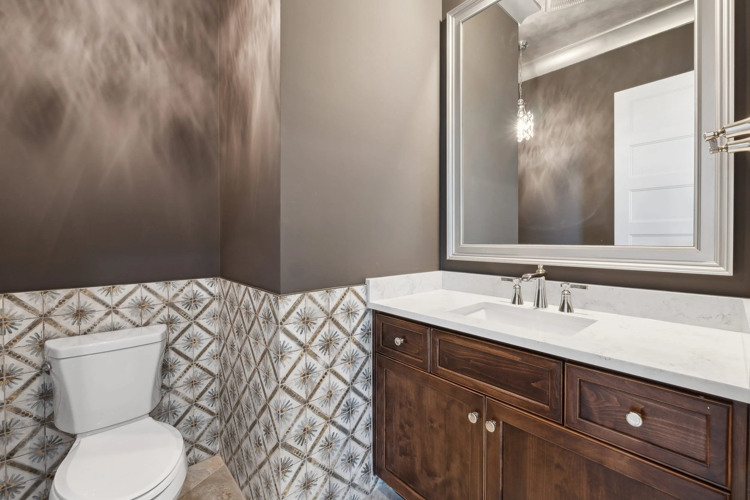 A bathroom with a toilet, patterned tile wall, and a wooden vanity with a white countertop and sink, large mirror, and a wall-mounted faucet. The wall above the tile is painted gray, and there is a hanging light fixture reflected in the mirror.