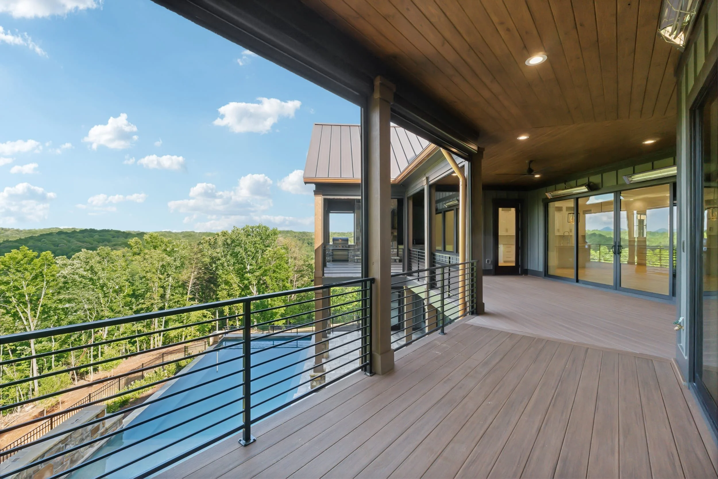 Balcony with wooden flooring, black metal railing, large glass doors, and recessed ceiling lights overlooking a green forested landscape with a bright blue sky and scattered clouds.