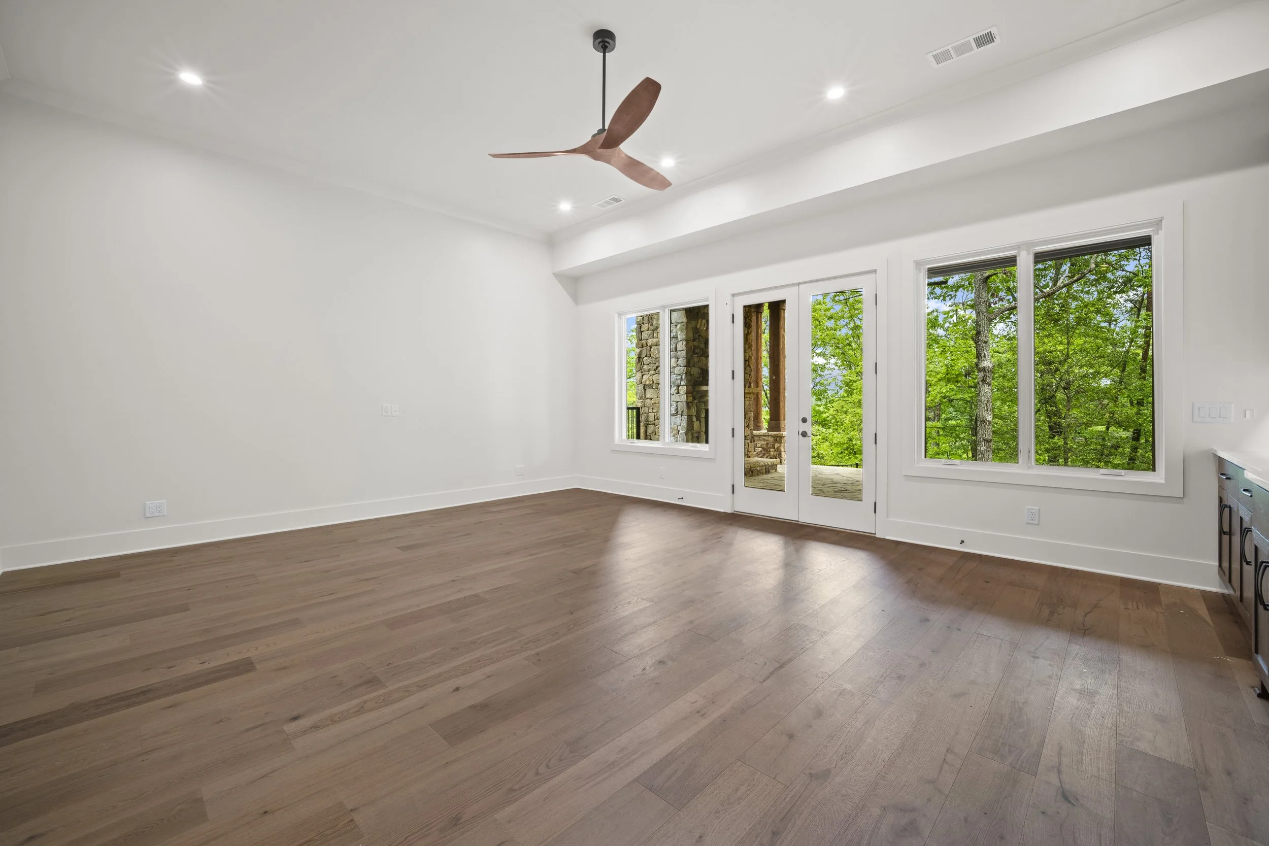 Empty living room with hardwood flooring, white walls, large windows showing green trees outside, and a ceiling fan.