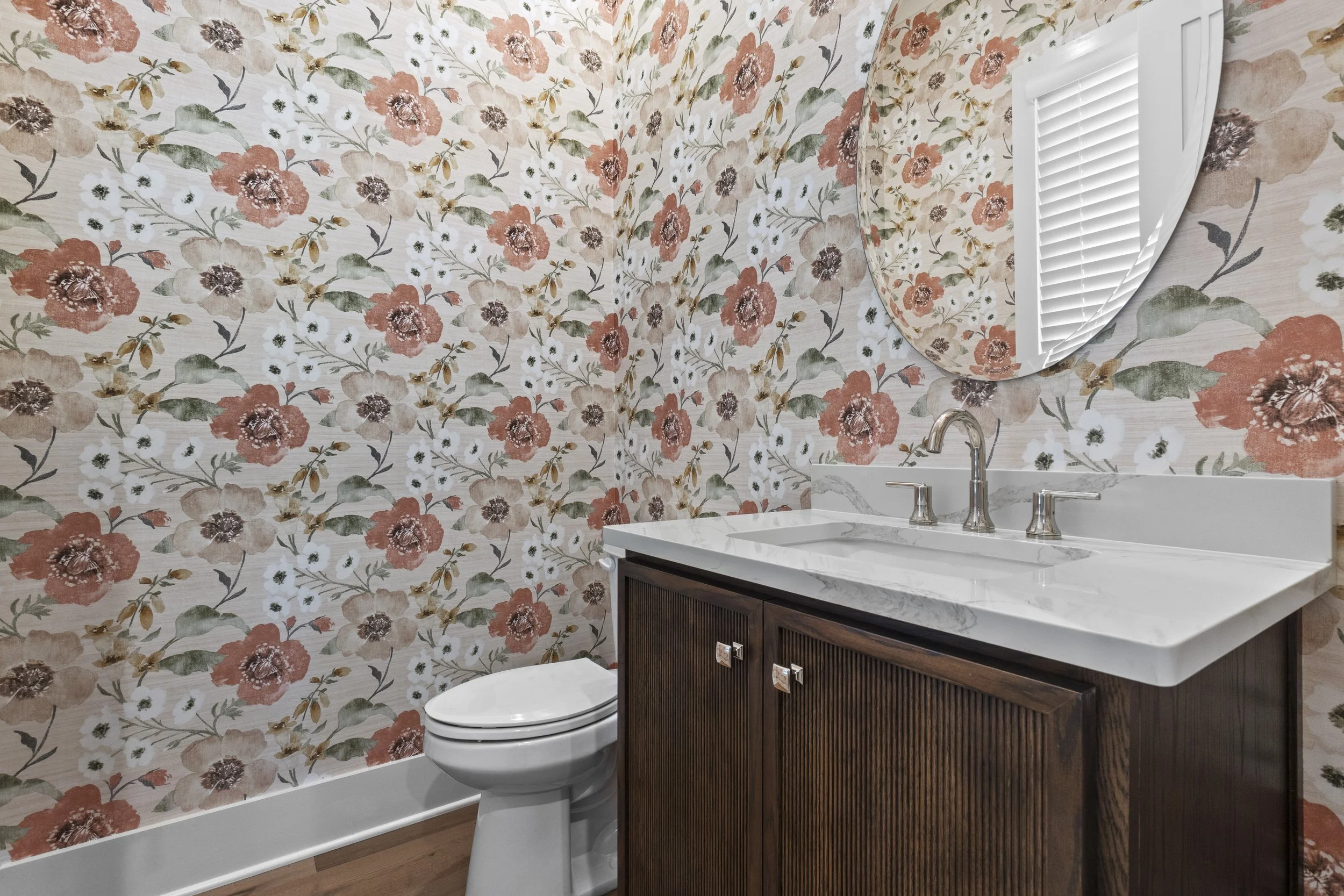 A bathroom featuring floral wallpaper with large pink, white, and brown flowers and green leaves. There is a white toilet and a dark wooden vanity with a marble countertop, a sink, and a metallic faucet. A window with white blinds is partially visibl