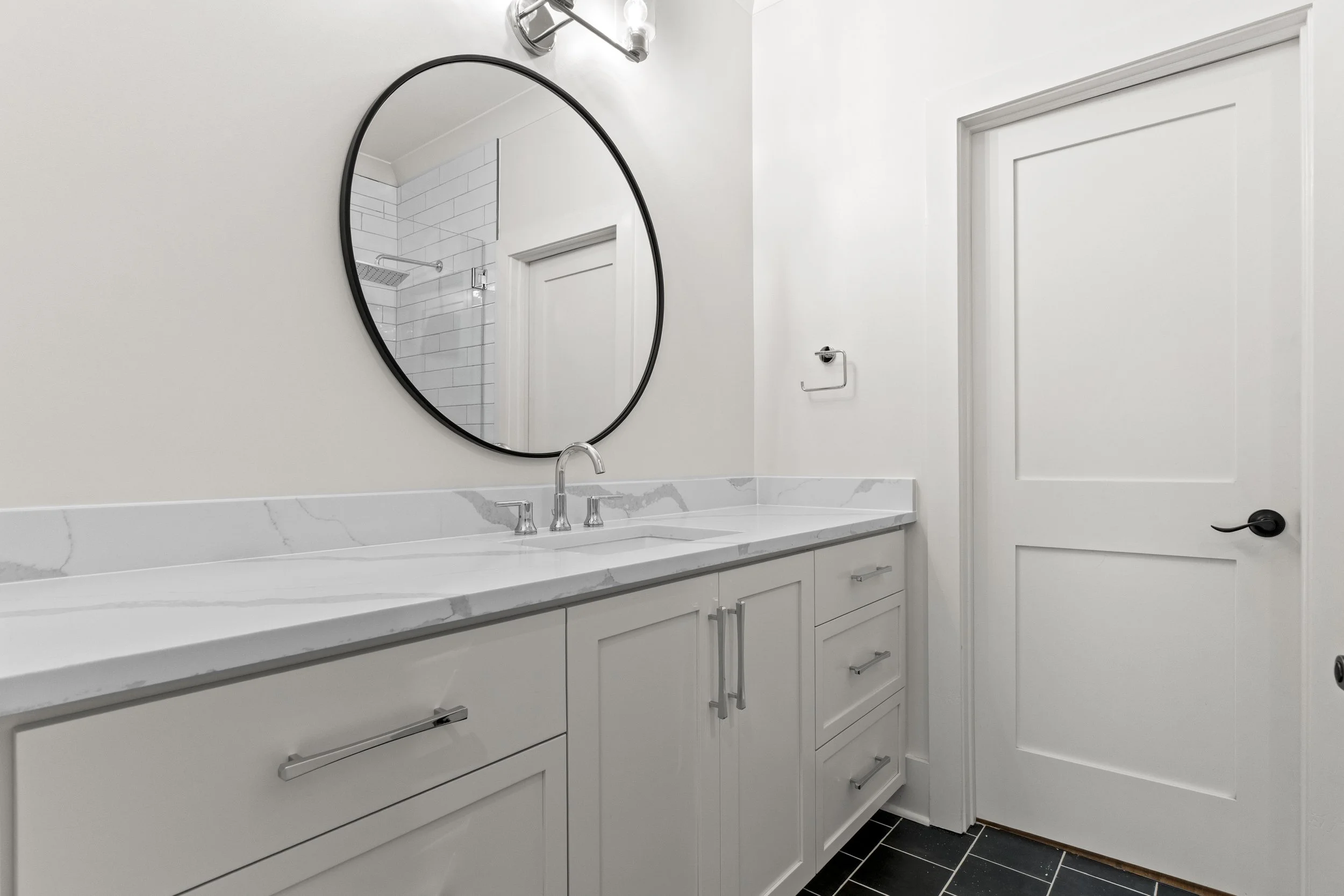 Modern bathroom with white cabinetry, a marble countertop, a round mirror, a faucet, and a partial view of a shower with white subway tiles and a rainfall showerhead.