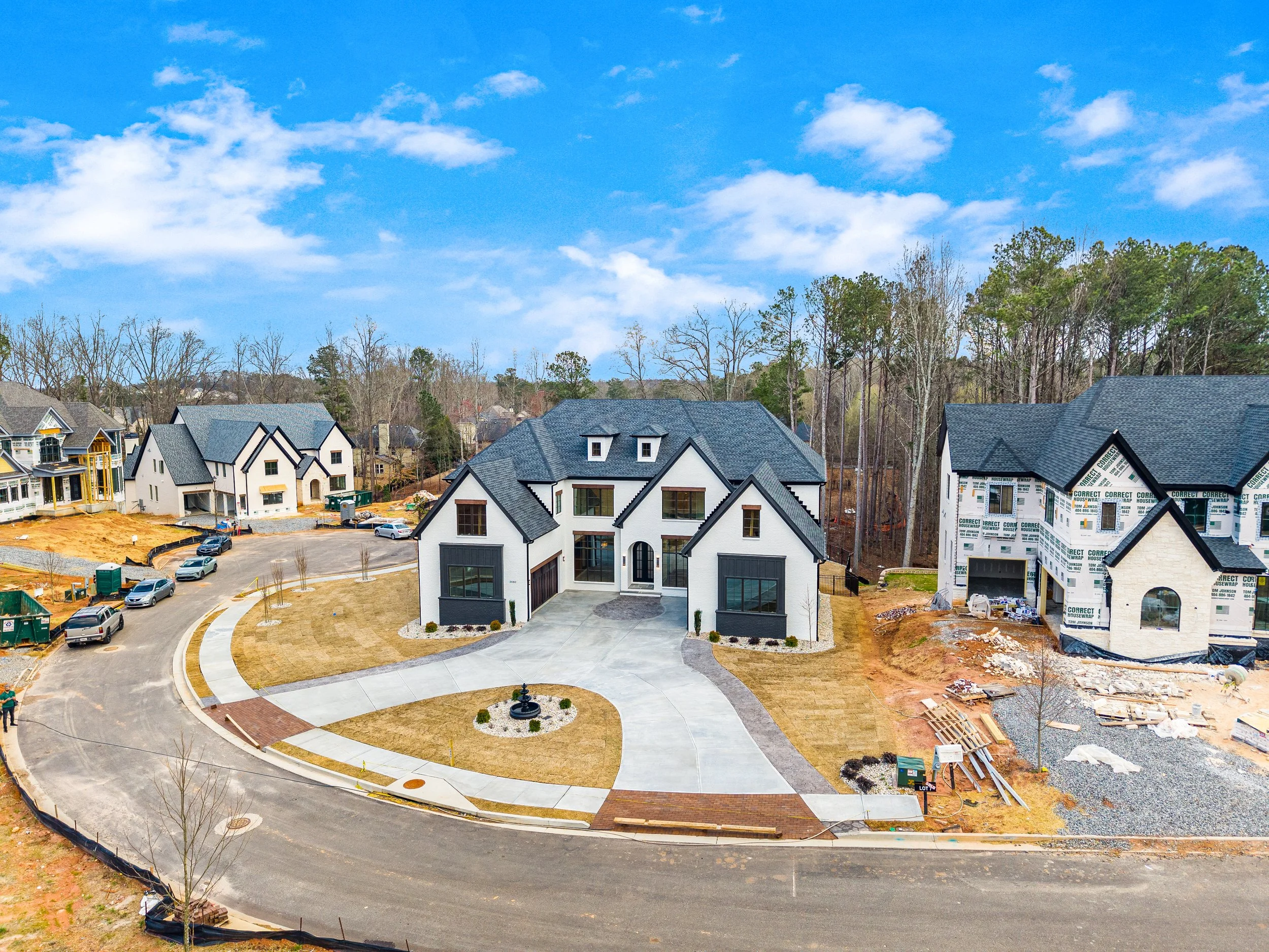 Suburban neighborhood under construction with several houses in various stages of completion, clear blue sky with scattered clouds, and a landscaped area with a fountain in the center.